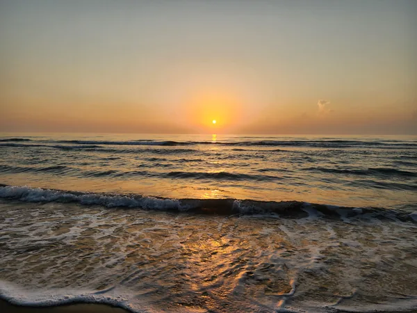 A low orange sun reflects across rolling waves and foamy surf at the shoreline.