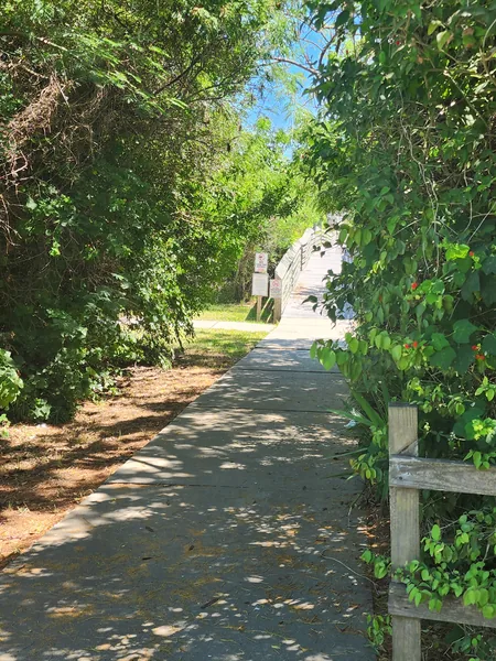 A shaded paved path leads through leafy vegetation toward the nature trail's wooden boardwalk.