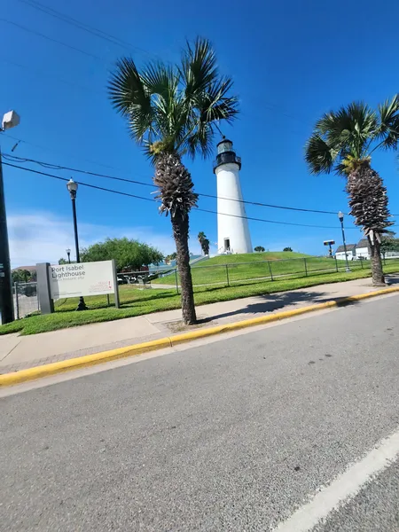 A Port Isabel Lighthouse sign at the foot of the hill, with the lighthouse and palm trees behind.