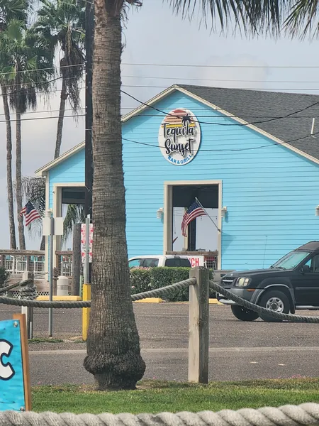 The sky-blue Tequila Sunset building with its round sign, seen past a palm trunk and a rope fence.