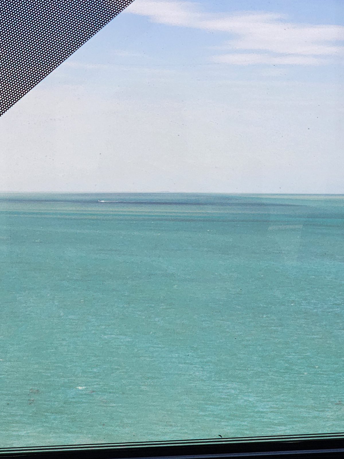 Turquoise bay water seen through a car window with a dotted tint edge, stretching to a hazy horizon where a tiny distant boat leaves a faint wake.
