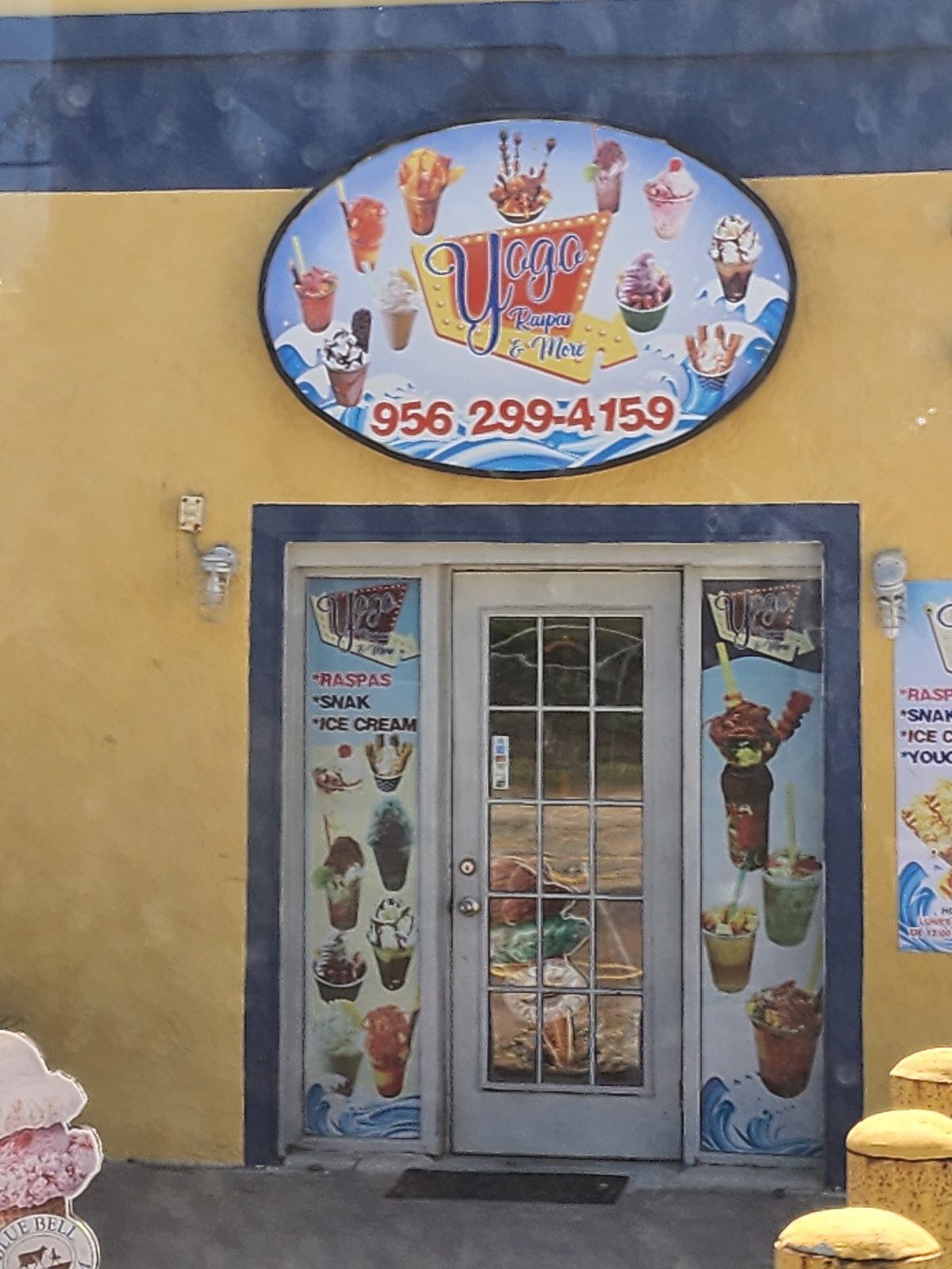 Oval Yogo Raspas & More sign with frozen-treat pictures and a phone number above a white glass door on a yellow storefront, flanked by dessert panels.