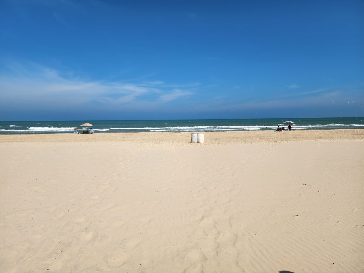 A broad sandy beach and rolling Gulf waves under a blue sky, with distant beachgoers.