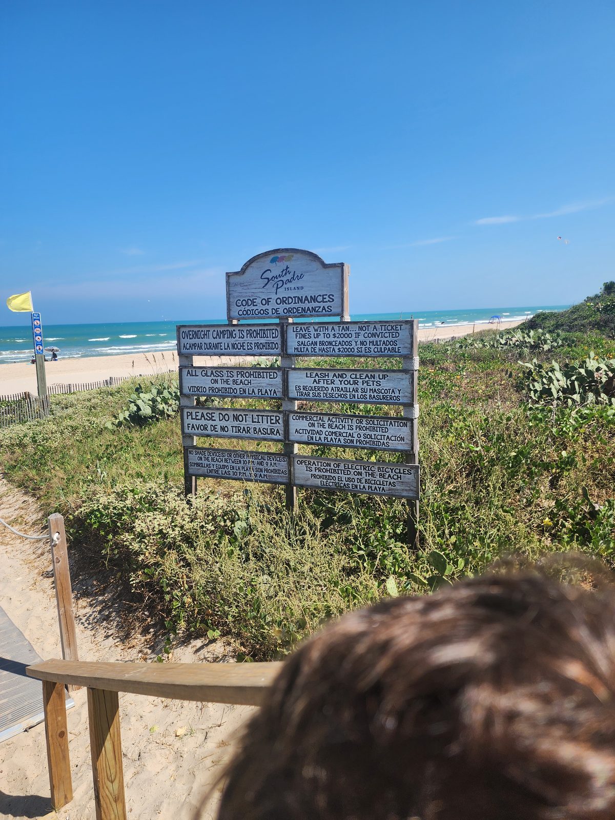 Bilingual Code of Ordinances sign beside a boardwalk railing, yellow flag and Gulf beyond, with the back of a person's head in the lower corner.