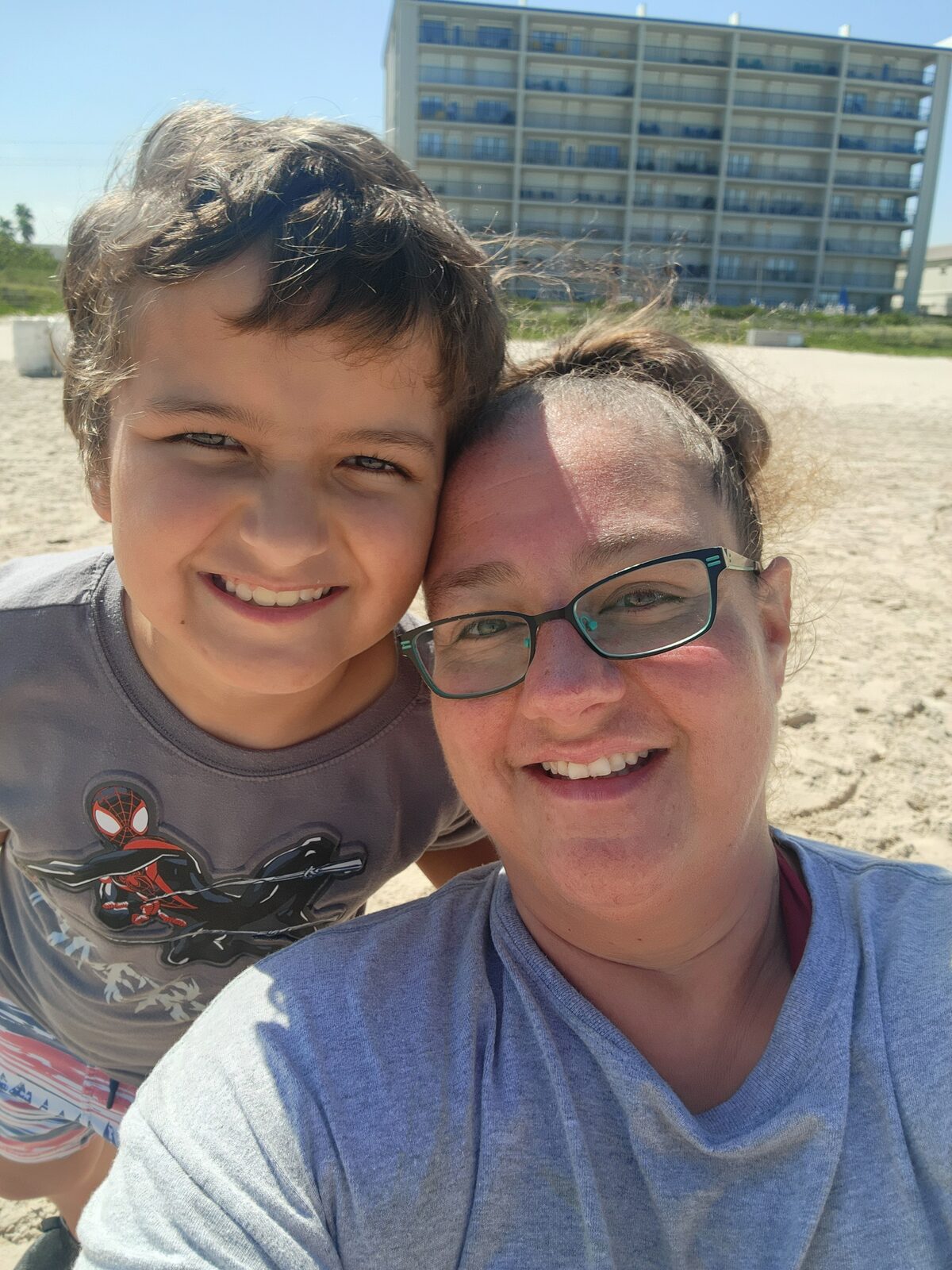 A smiling adult and child share a cheek-to-cheek beach selfie in bright sun, with dune grass and a tall beachfront high-rise behind them.
