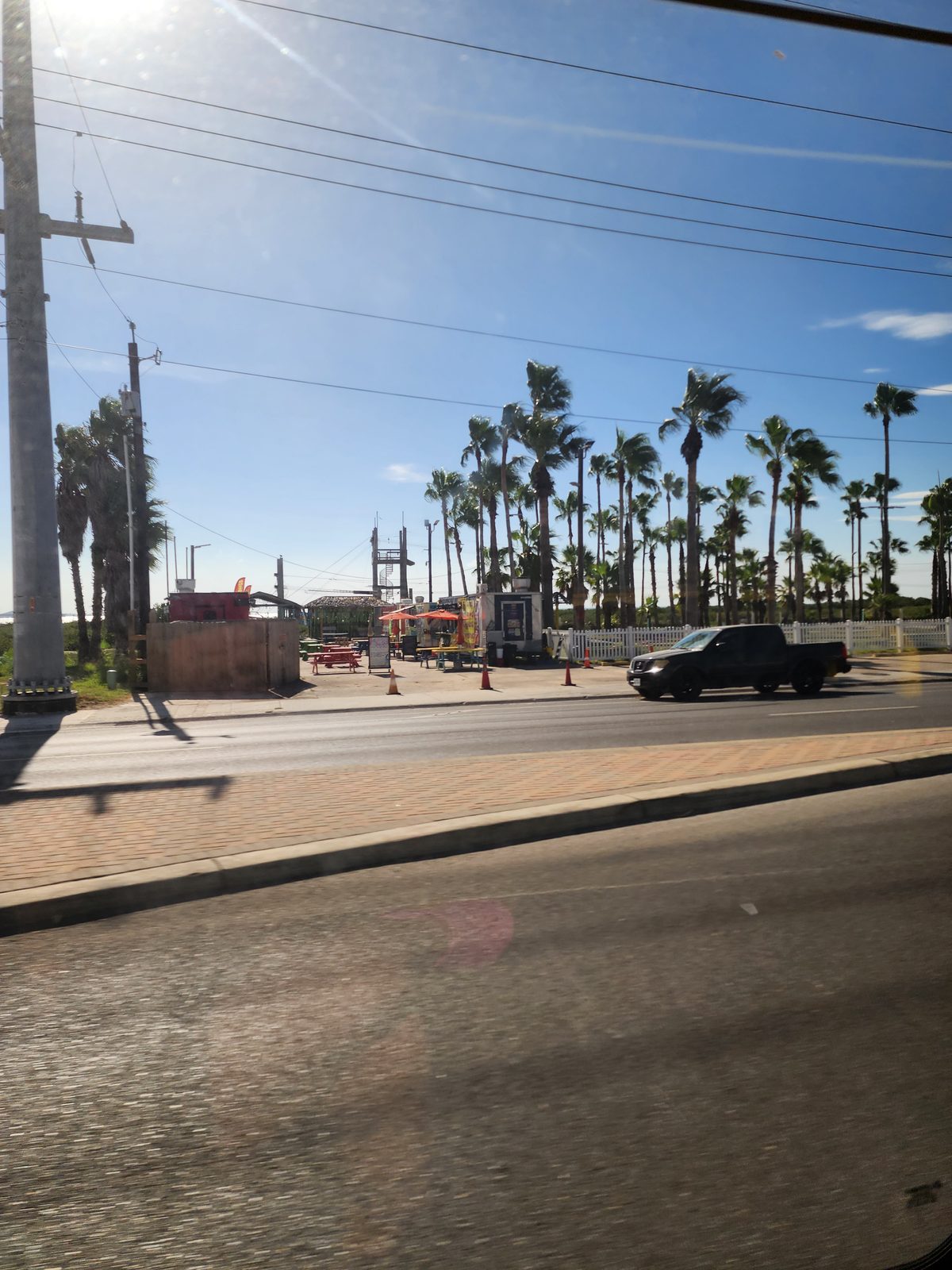 Wide road with a brick median and a passing pickup, and across the street an open-air lot with food trailers, colorful picnic tables and tall palms.