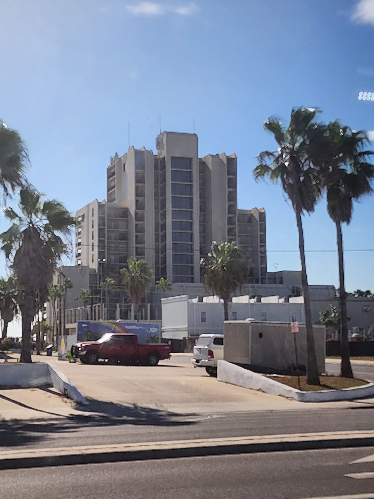 Tall stepped concrete high-rise tower with balconies rises behind palm trees, low buildings and two parked pickup trucks across a road.