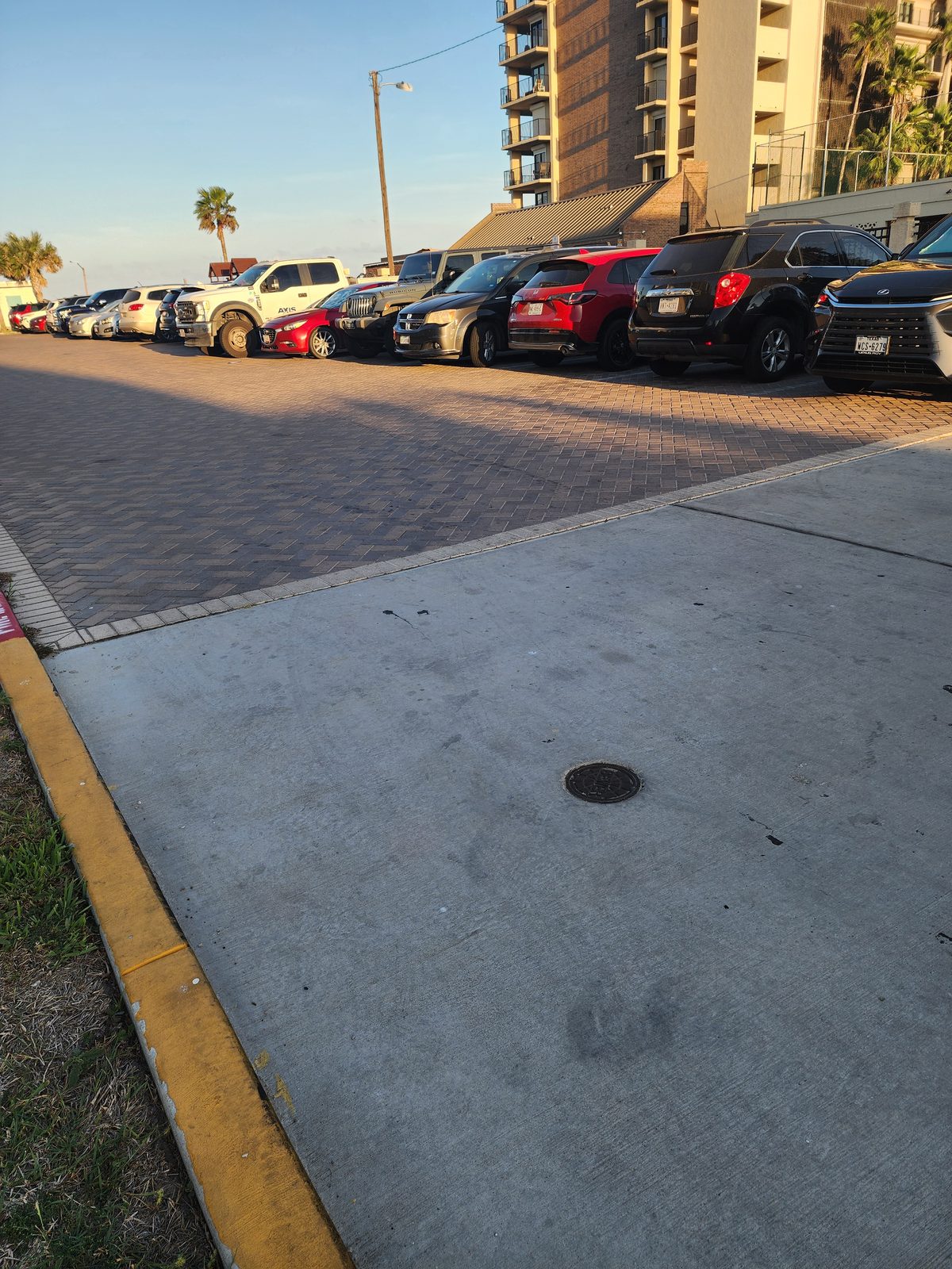 Row of parked cars and a pickup in a brick-paver parking lot beside a tall high-rise with balconies and palm trees in low evening light