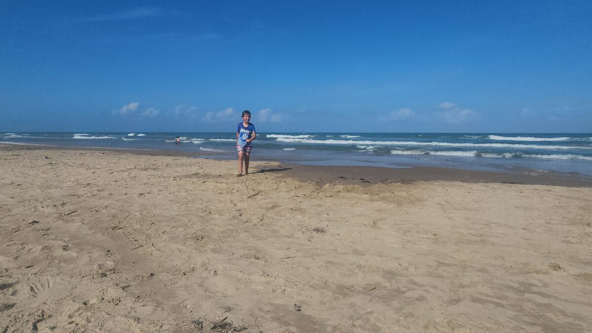 A child stands on a broad beach beside shallow surf.
