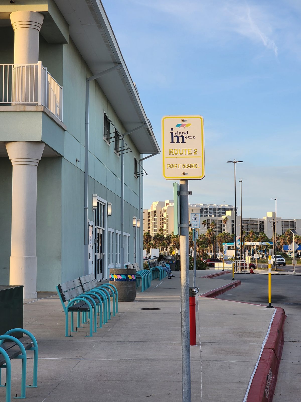 Island Metro Route 2 Port Isabel stop sign beside teal benches along a mint-green two-story building, with condo towers behind.