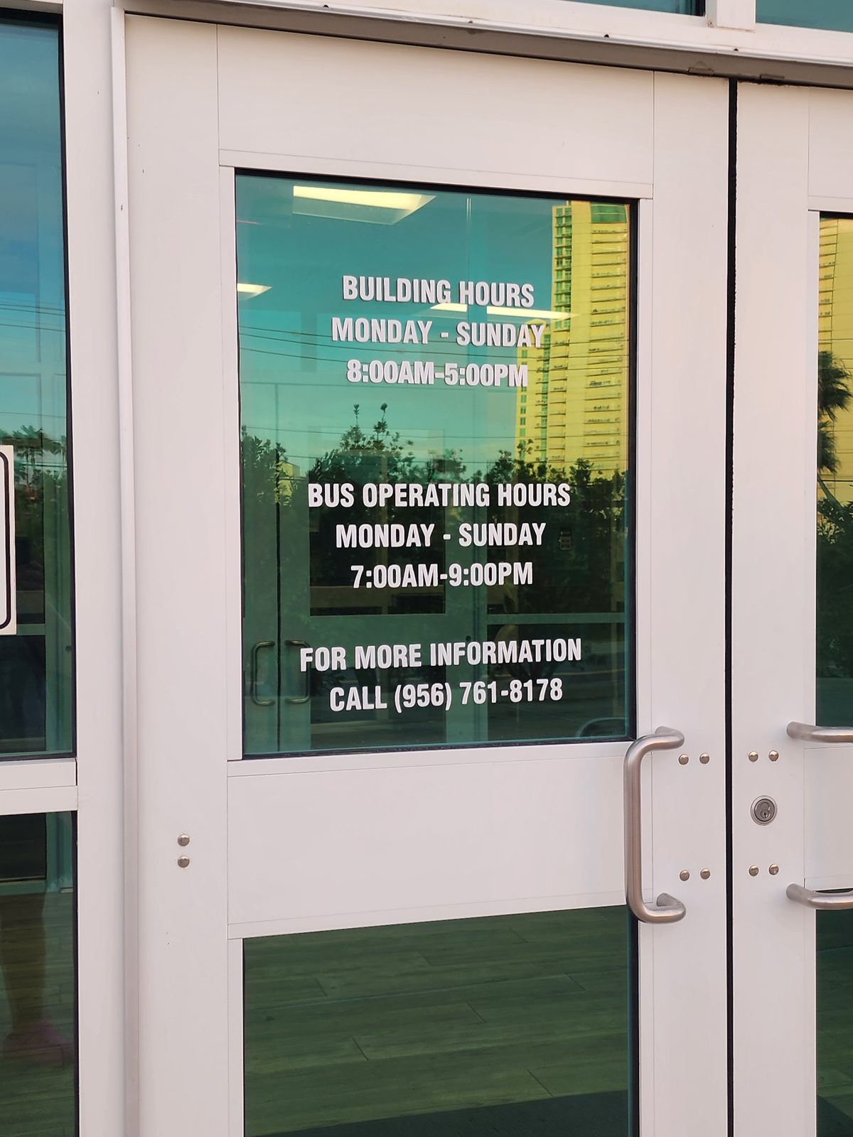 Glass entrance door lettered with building hours of 8 AM-5 PM and bus operating hours of 7 AM-9 PM daily, reflecting a tall high-rise tower and palm trees.