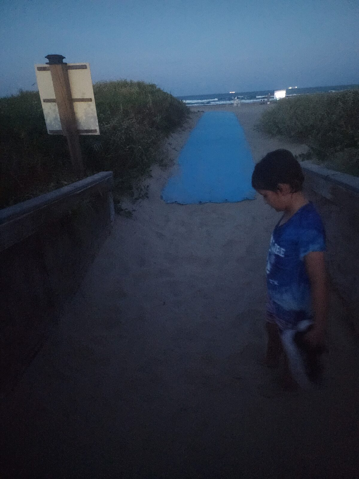 A child stands looking down on a sandy beach-access path through the dunes at dusk, with a blue mobility mat leading toward the Gulf ahead.
