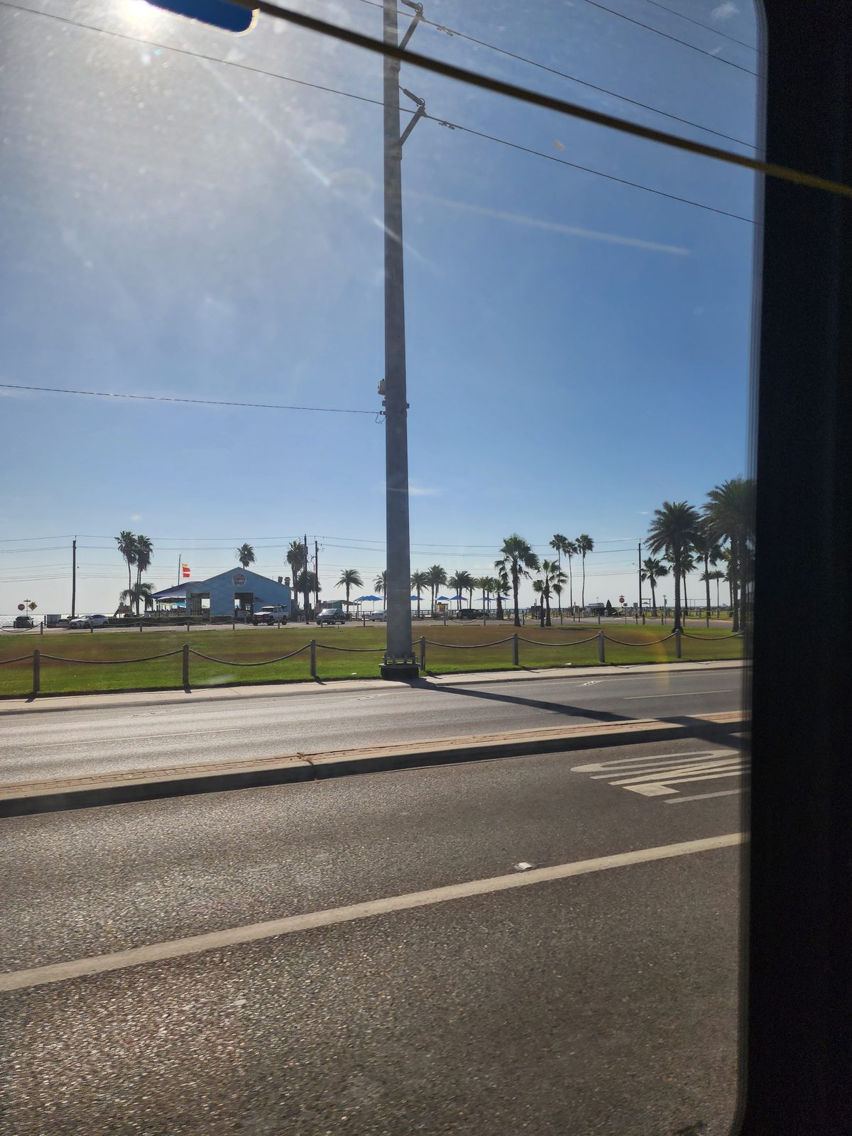 A wide road runs past a mowed lawn behind a rope fence, a utility pole, parked cars, a low blue building with umbrellas and palms, water on the horizon.