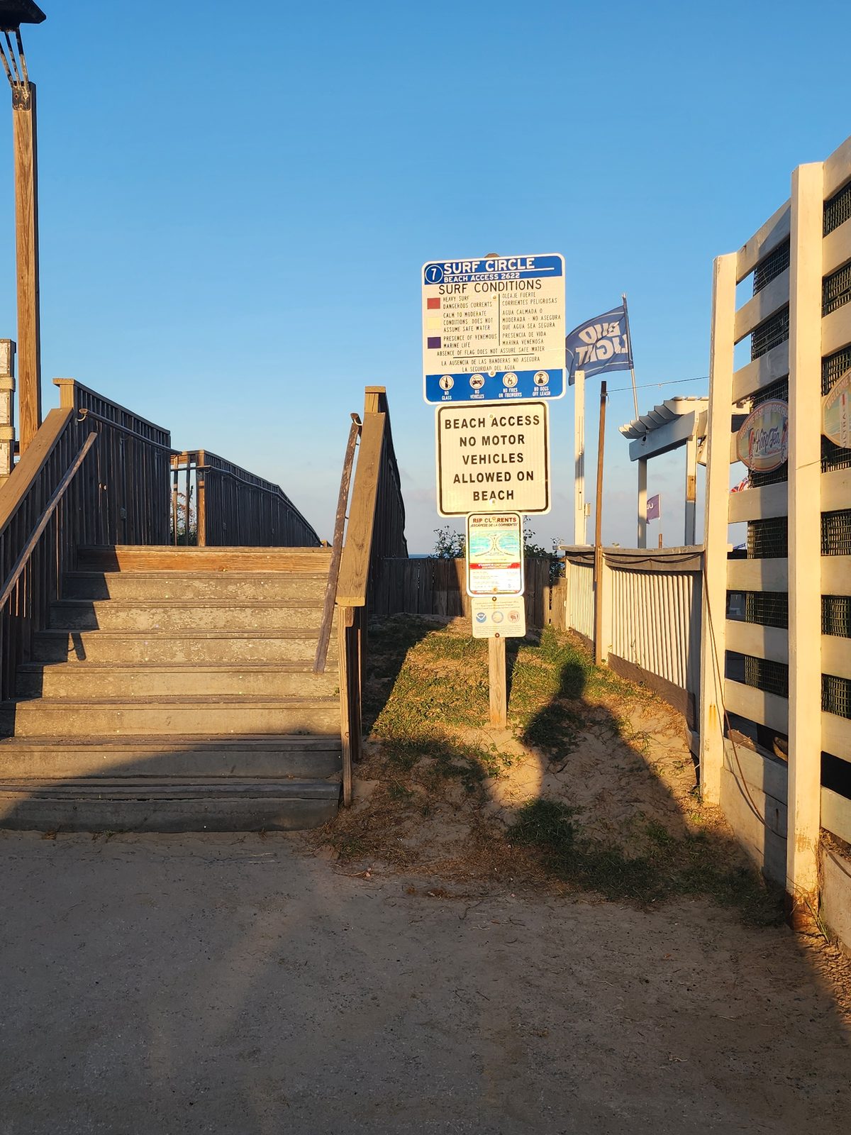 Surf Circle Beach Access 7 sign with a surf-conditions flag legend, a no-motor-vehicles notice and a rip-current warning beside wooden stairs to the beach.
