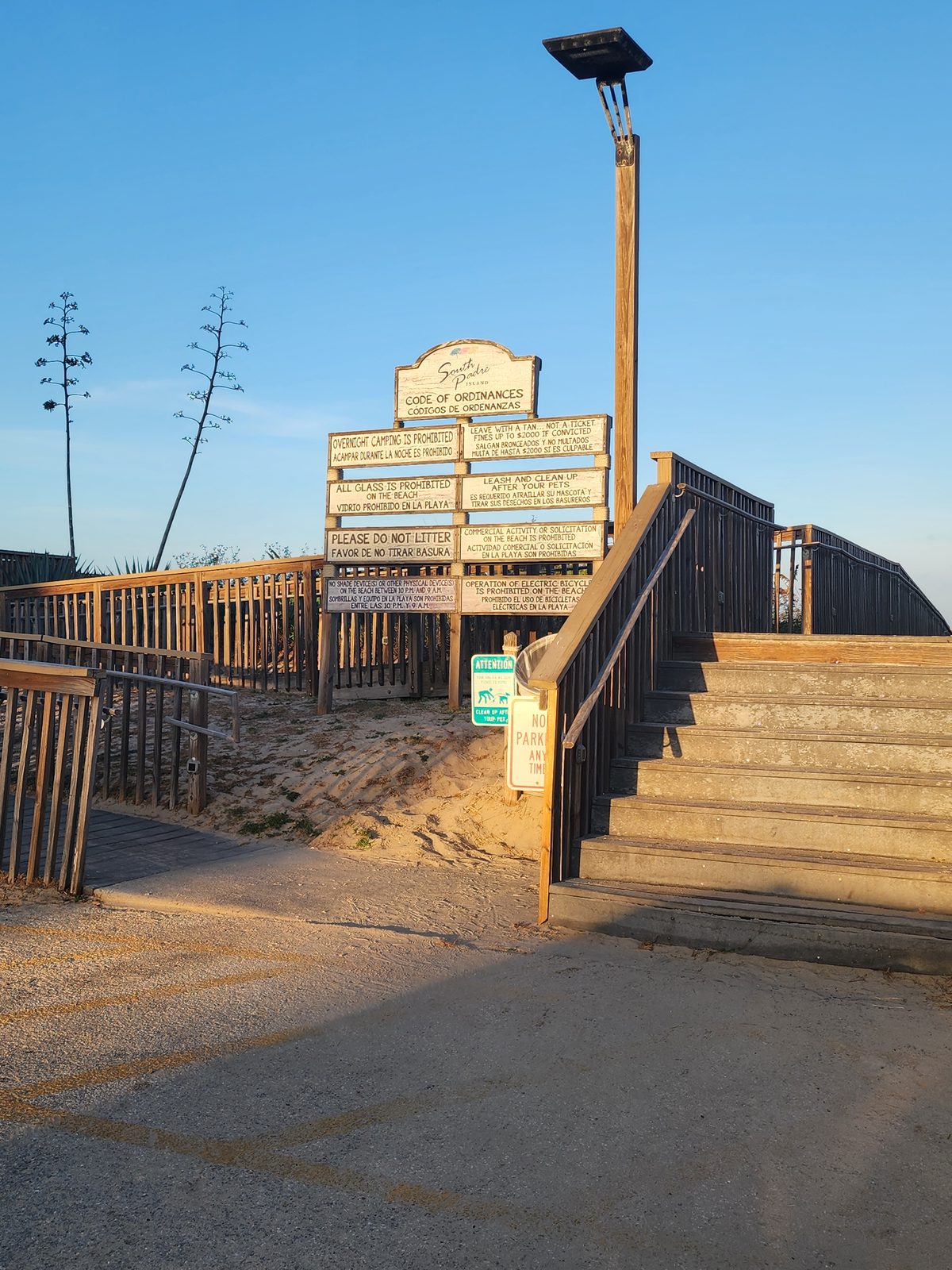 Bilingual South Padre Island code-of-ordinances sign board beside wooden walkover stairs and a ramp, lit by low evening sun.