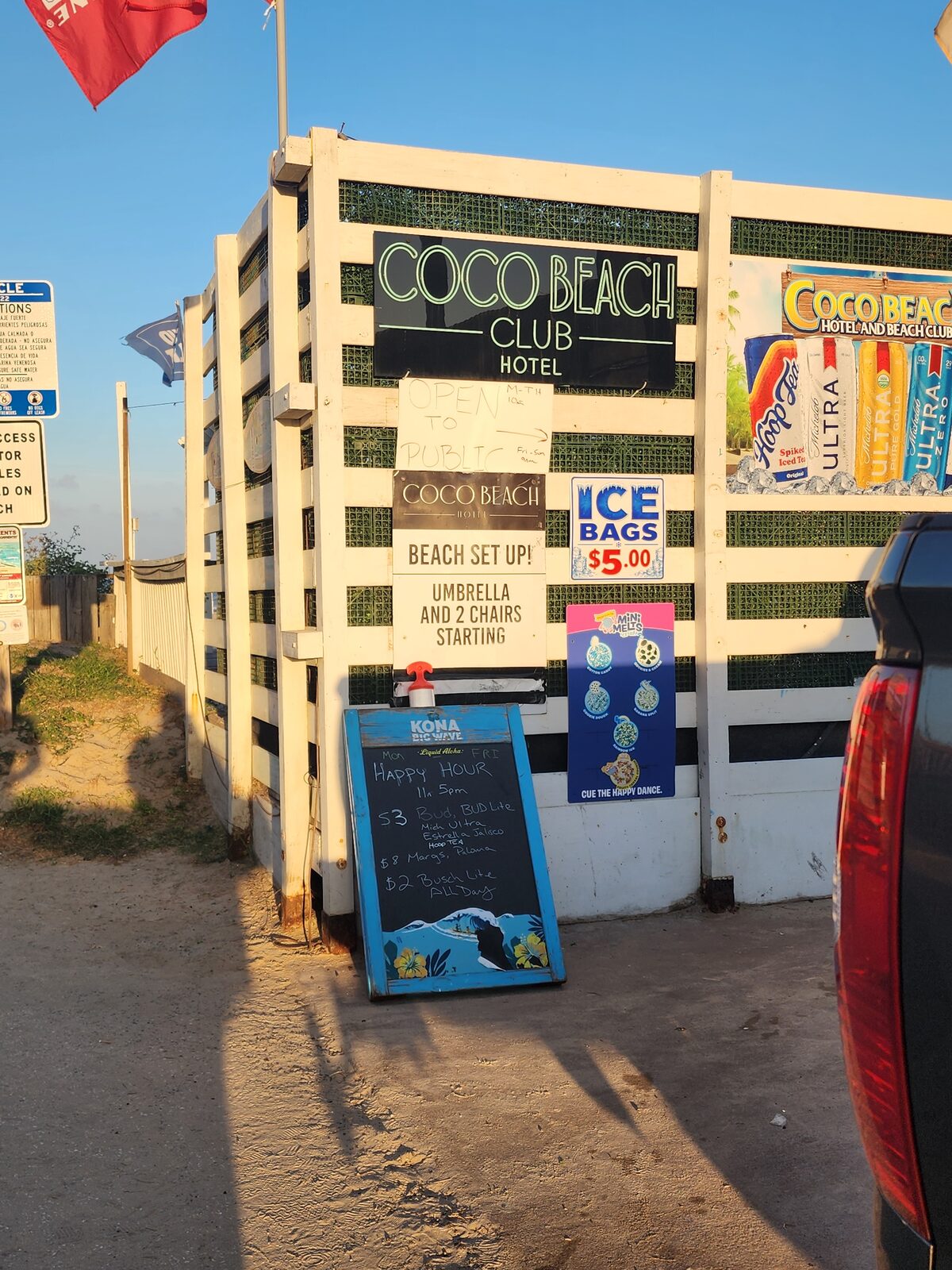 Coco Beach Club Hotel's signboard wall beside a beach access, with beach setup, ice bag and happy-hour signs.