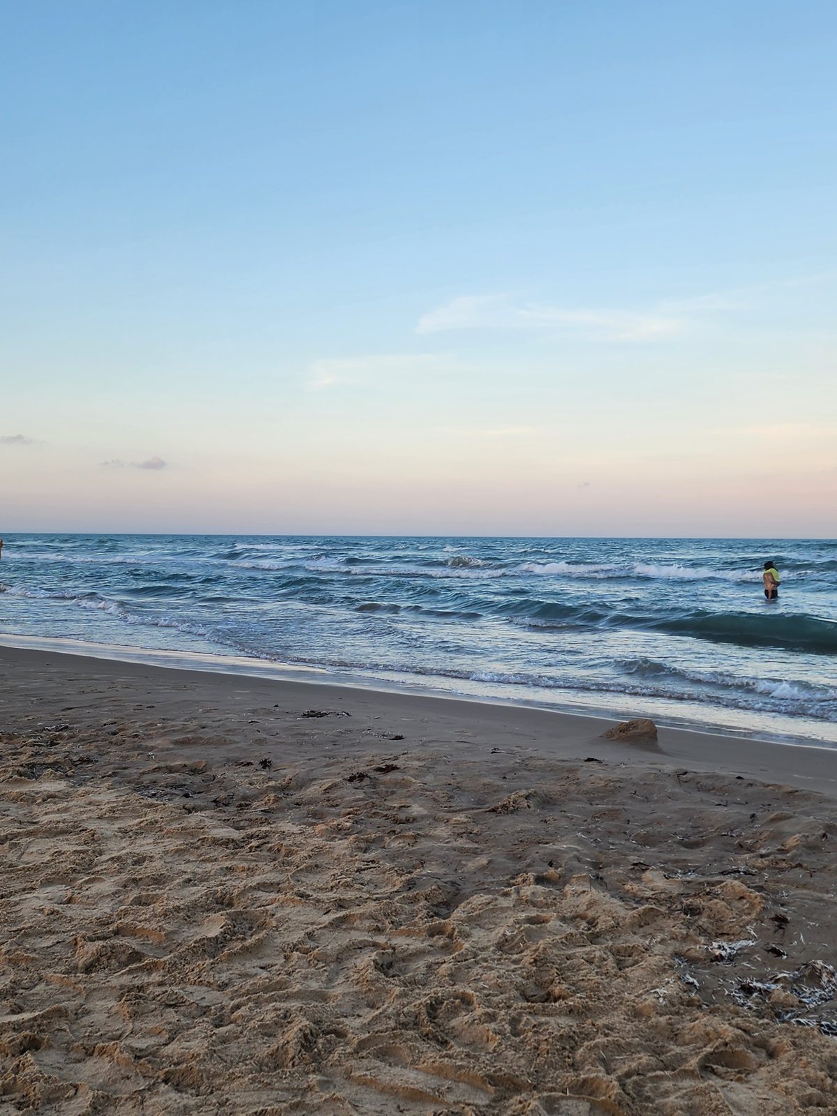 Small waves roll onto a wide, footprint-covered Gulf beach under a pale evening sky, with one person wading waist-deep in the surf.