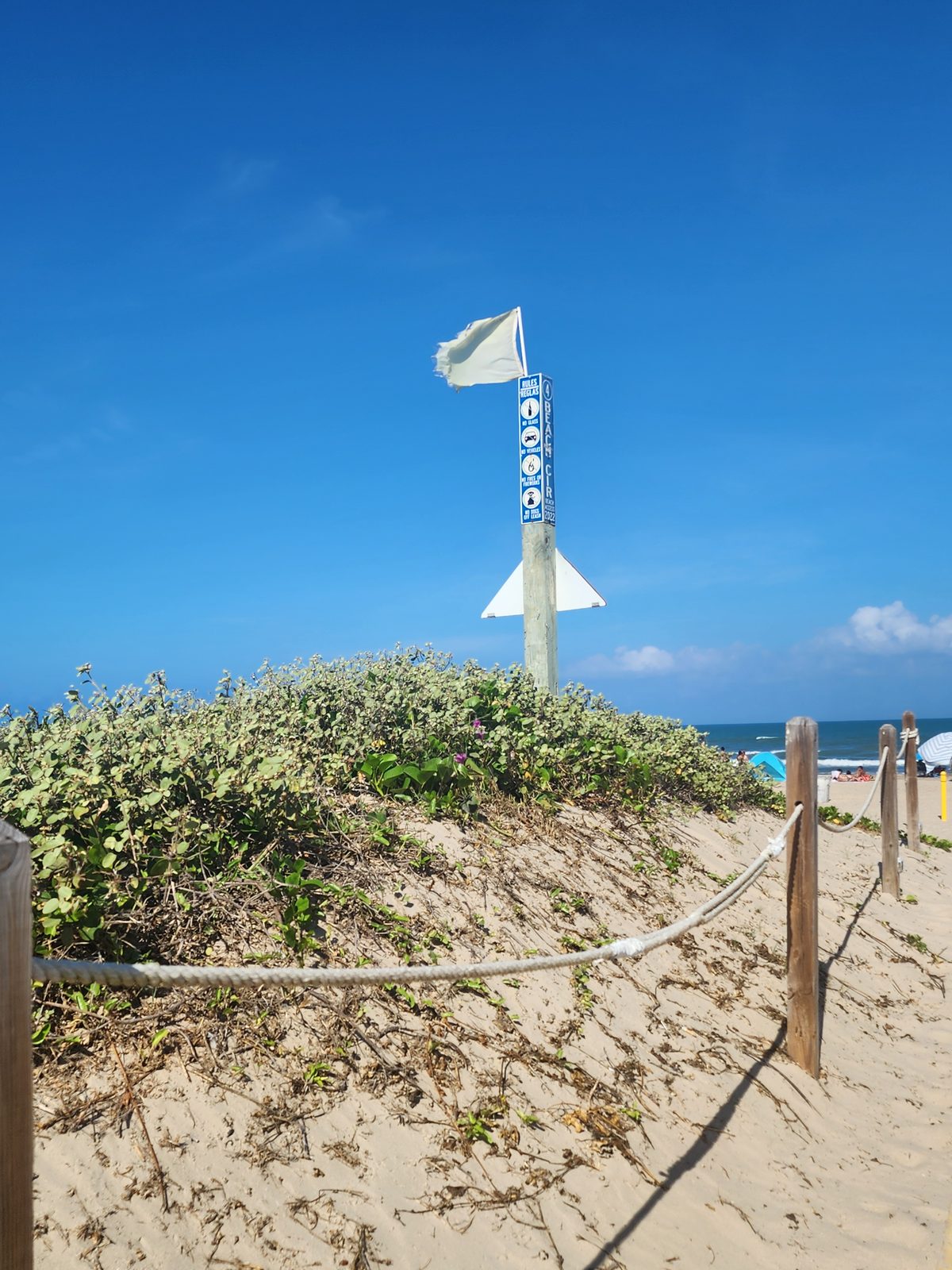 A roped dune path leads toward a beach beneath a white flag.
