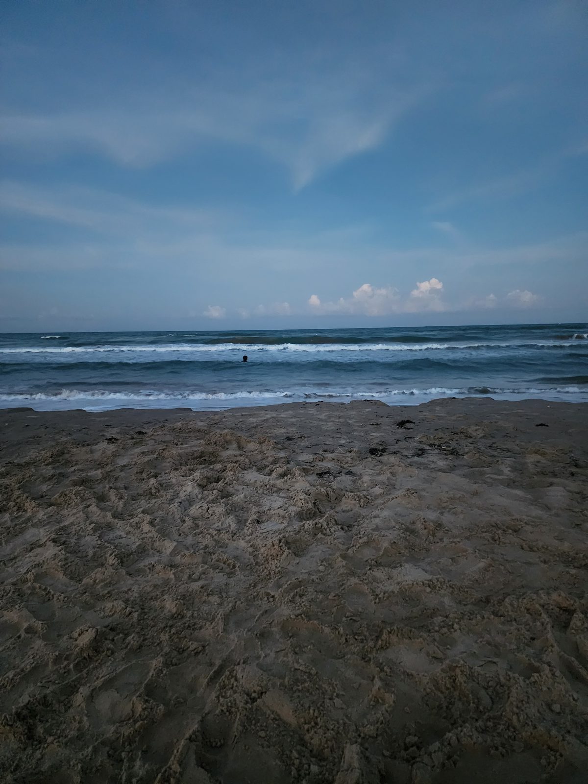 Wide sandy beach and lines of Gulf waves at dusk, with a single swimmer's head visible out past the breakers.