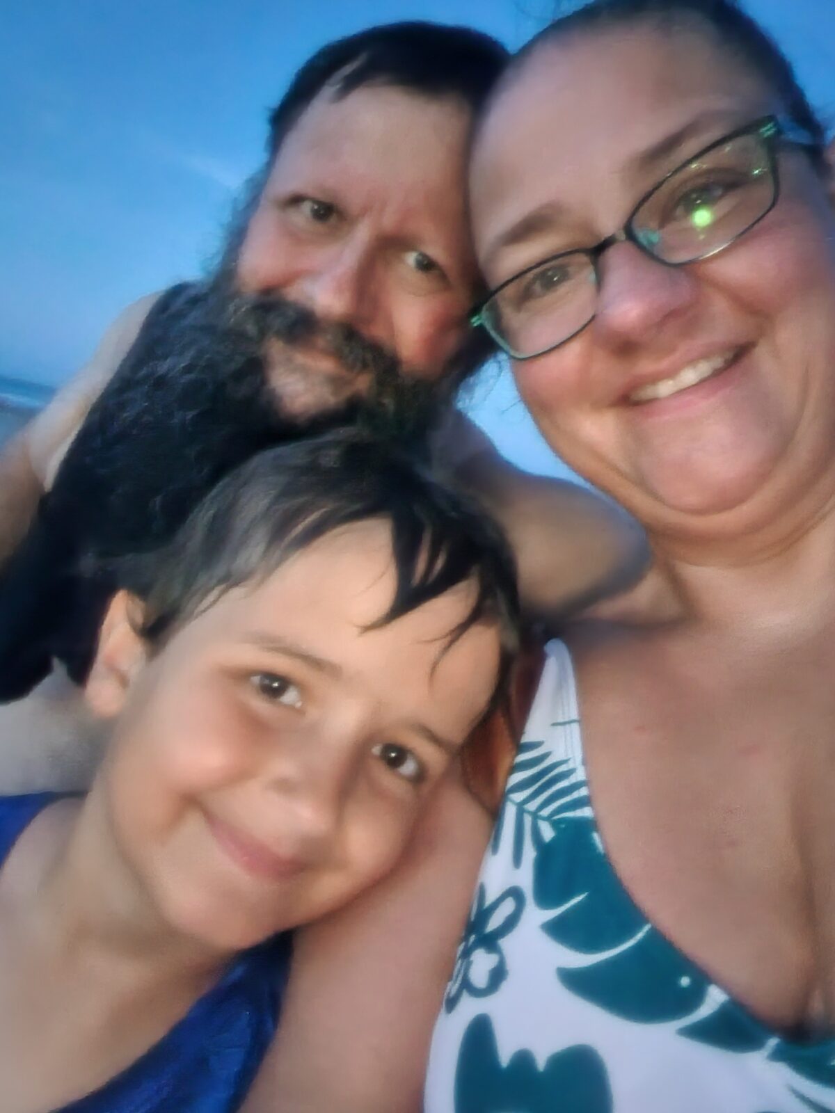 A close family selfie of two adults and a child on the beach at twilight.
