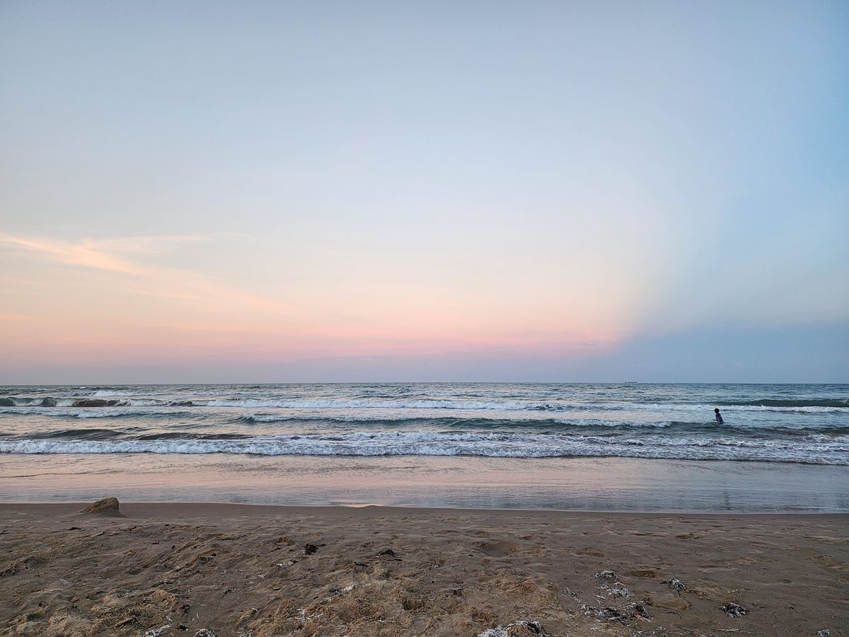 A lone person stands waist-deep in the Gulf surf beneath a wide pastel evening sky, with wet sand reflecting the colors.