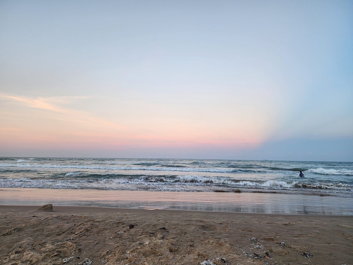 A lone figure wades waist-deep in Gulf surf under a pastel pink and blue evening sky over a wide sandy beach.