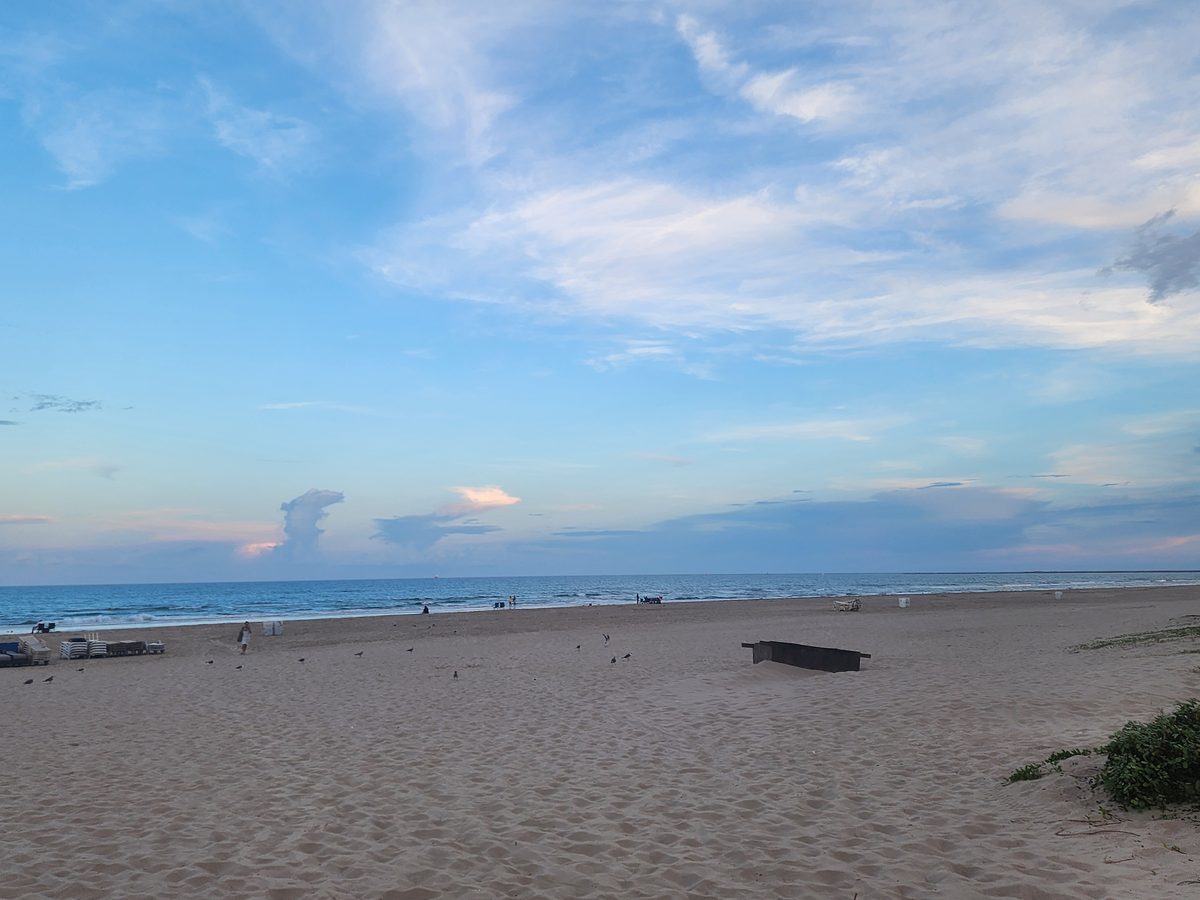 Distant visitors stand near the waterline on a sandy beach at sunset.