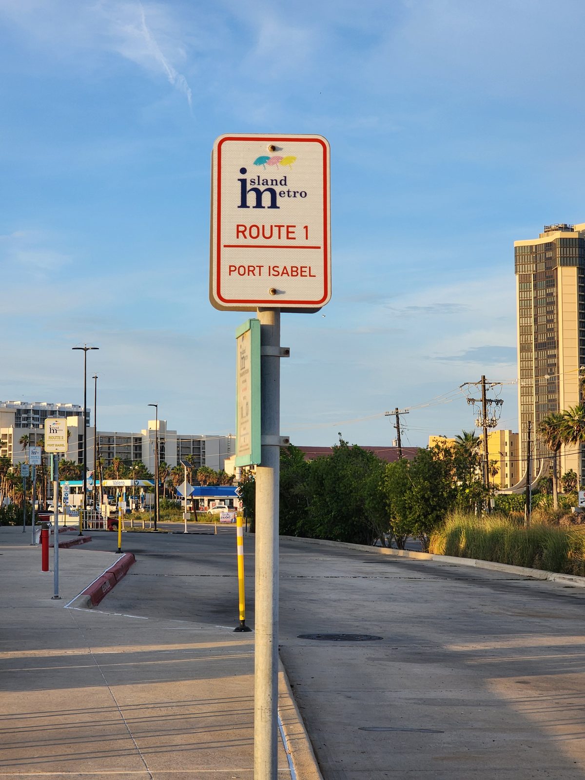 Island Metro Route 1 sign for Port Isabel on a pole in a parking lot, with a gas station and condo towers behind.