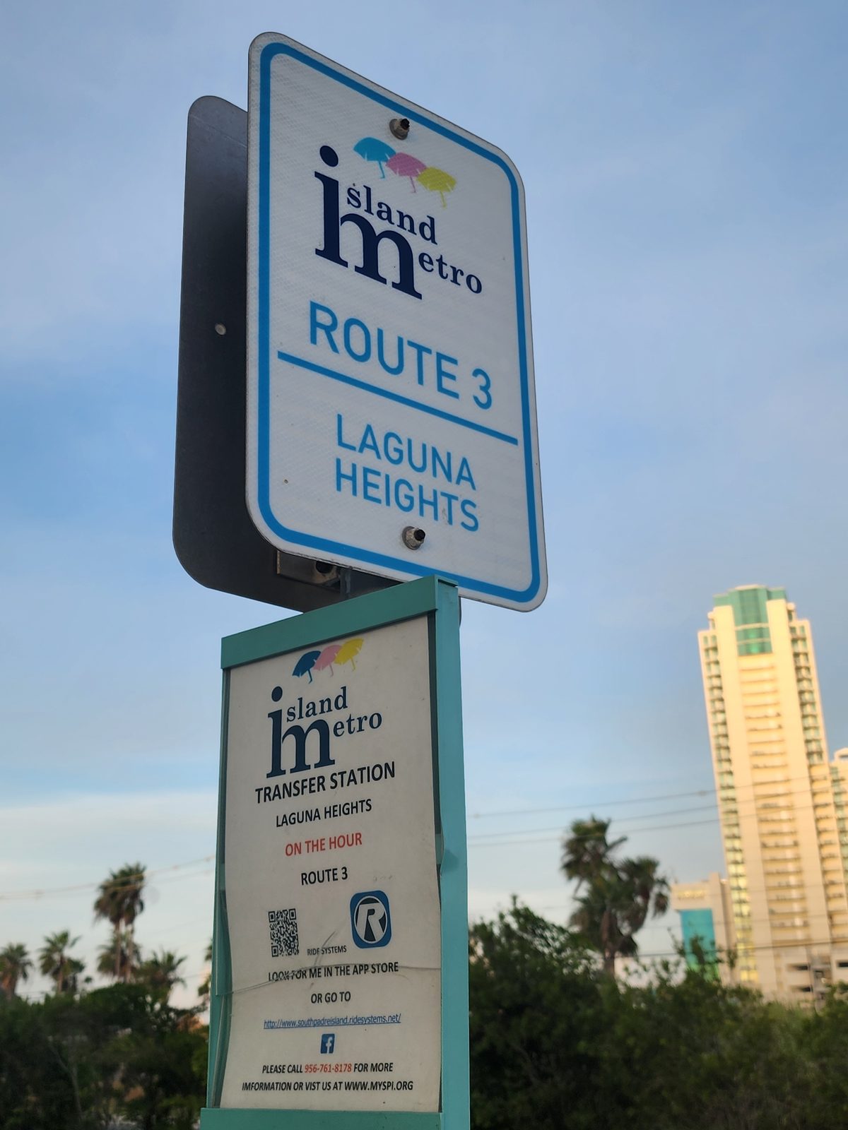 Island Metro Route 3 Laguna Heights sign above a transfer-station placard with a QR code, a tall condo tower and palms behind.