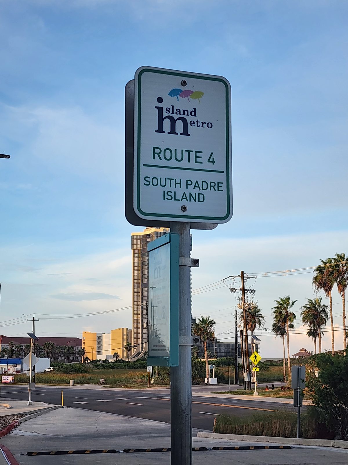 Island Metro Route 4 bus stop sign on a pole beside a road, with palm trees and a high-rise tower behind it in evening light