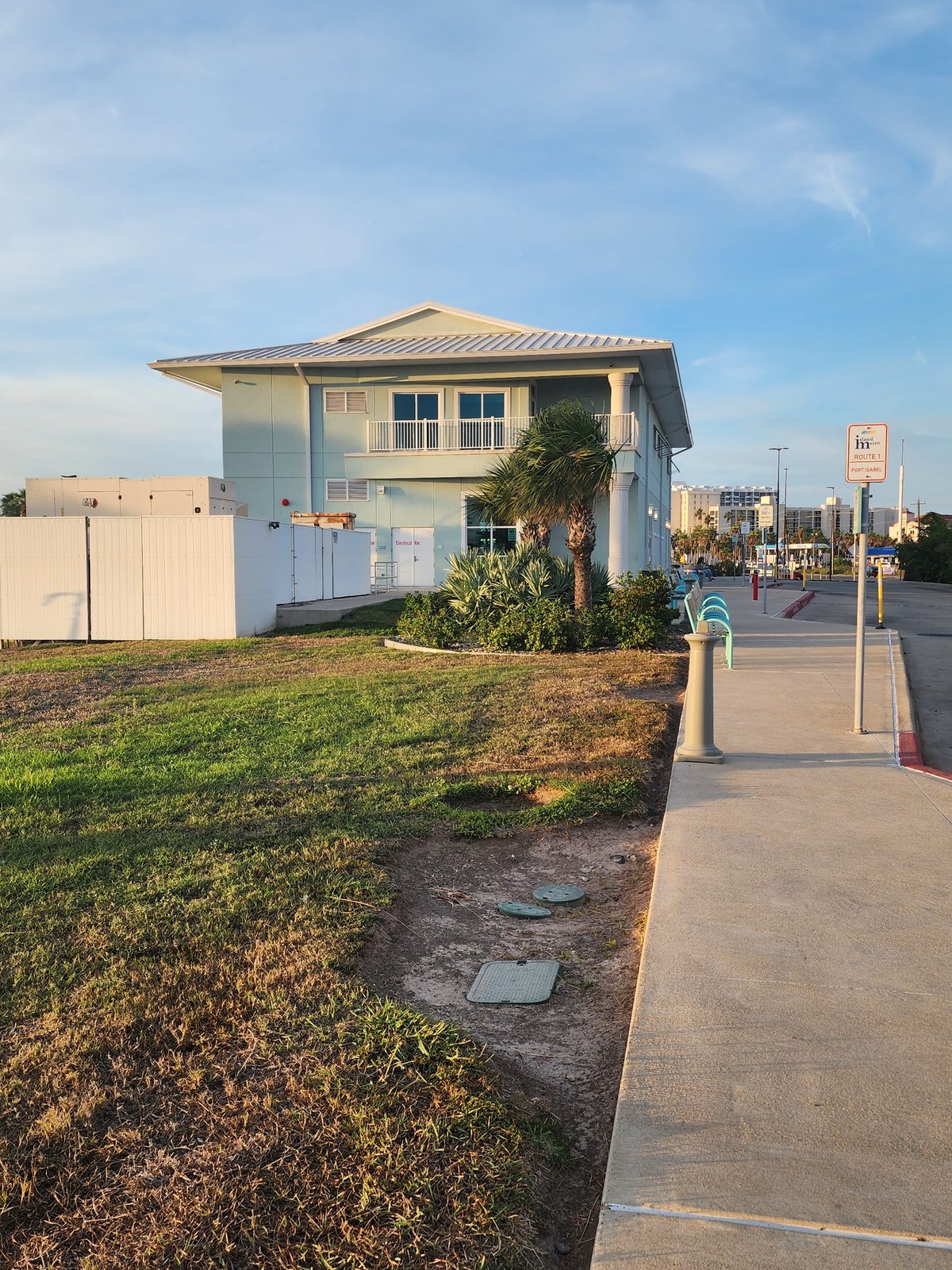 Two-story pale green building with white trim, a balcony and a metal roof, one palm tree, and a sidewalk with an Island Metro Route 1 sign.