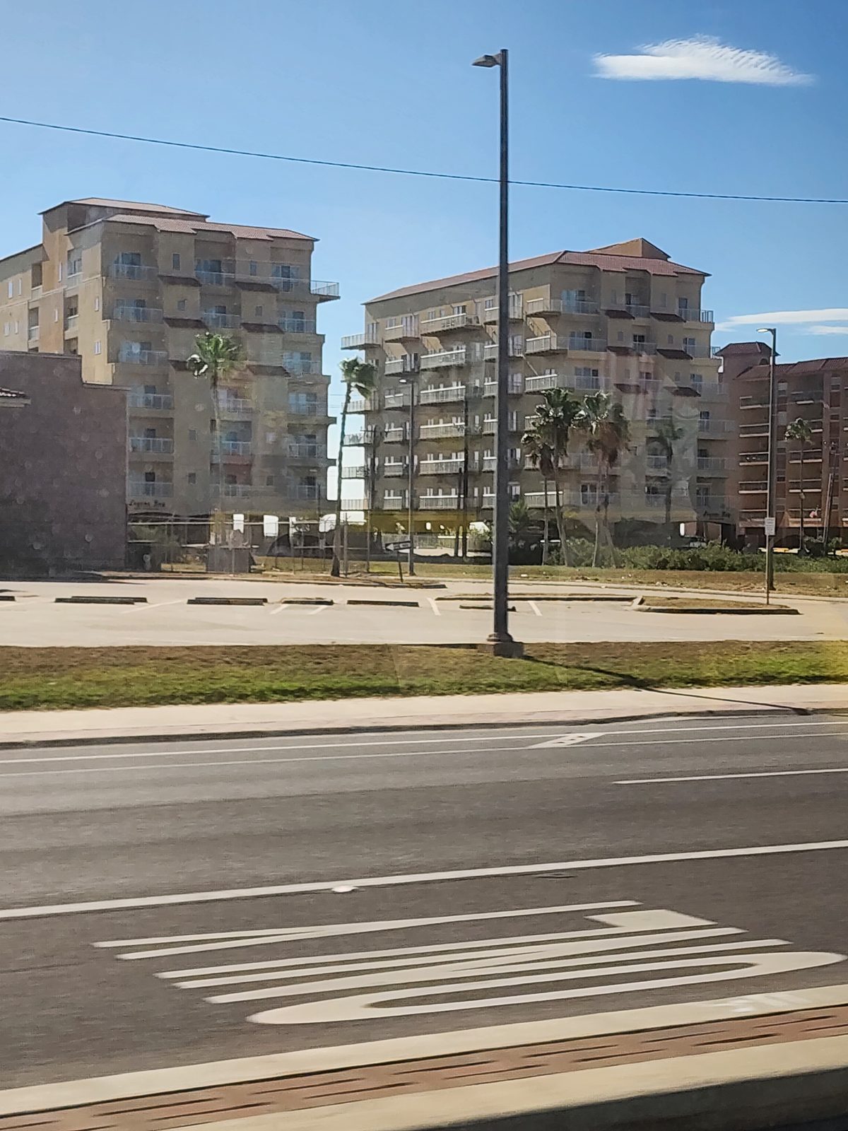 Two matching tan mid-rise buildings with white-railed balconies and tile roofs stand behind palms and an empty striped lot across a wide road.