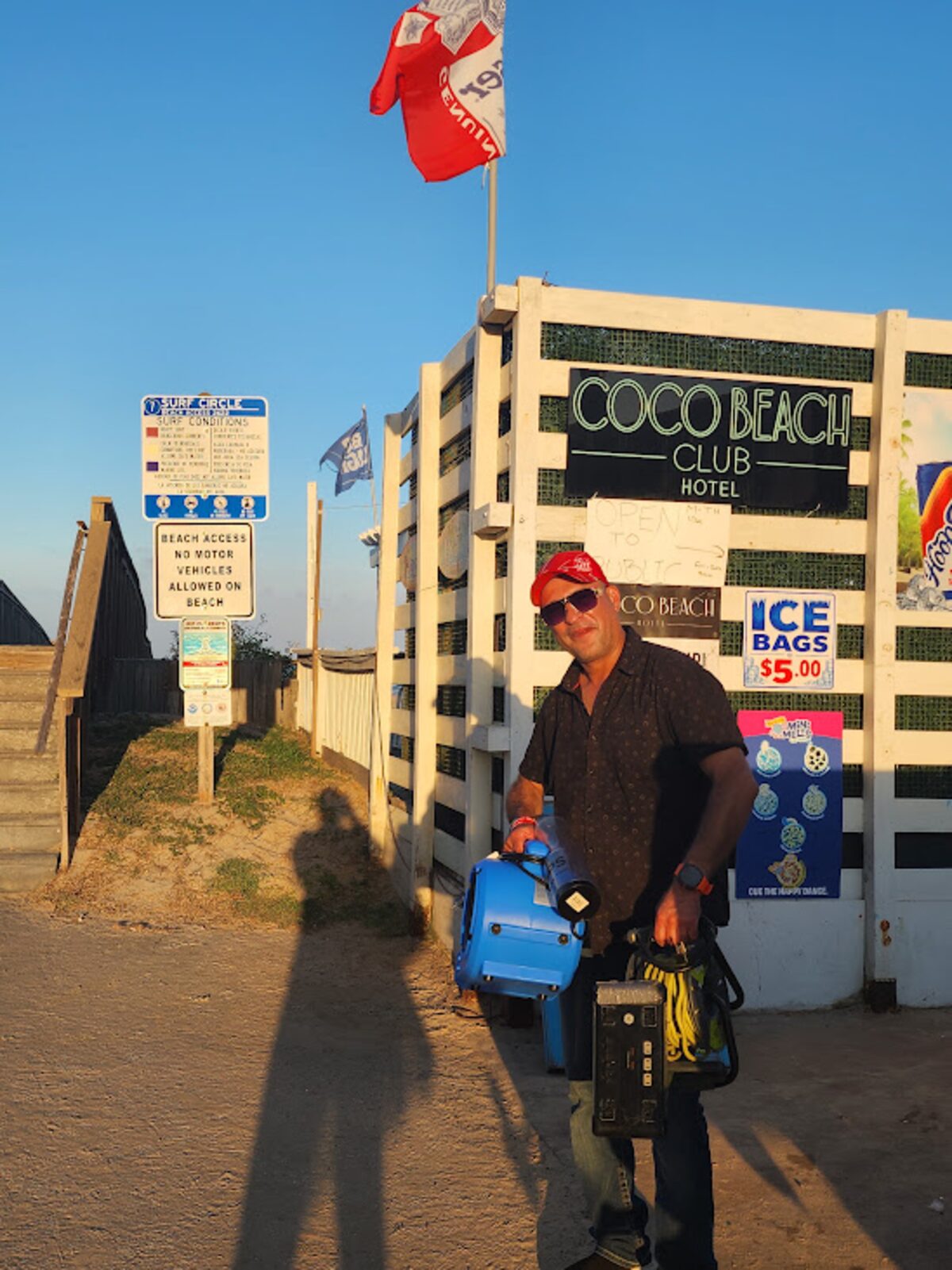 A smiling man in a red cap carries a blue portable fan and gear past the Coco Beach Club Hotel signs beside a beach access stairway.