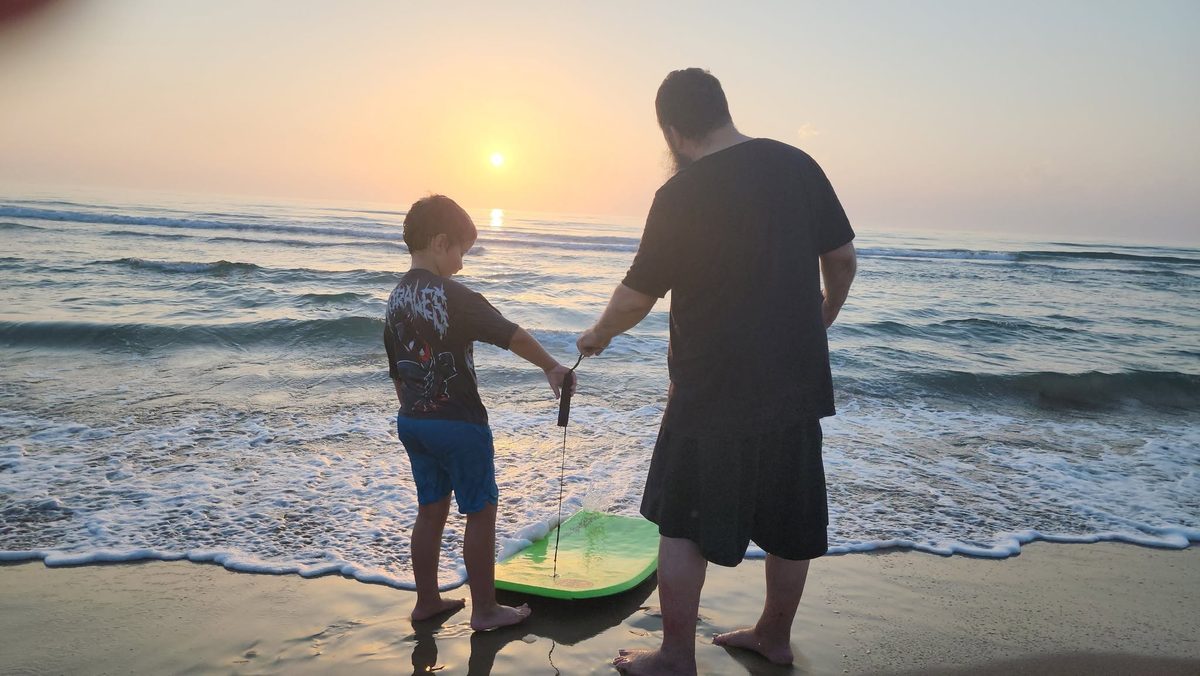 An adult and a child stand barefoot at the water's edge, holding the leash of a green board as the sun rises over the Gulf.