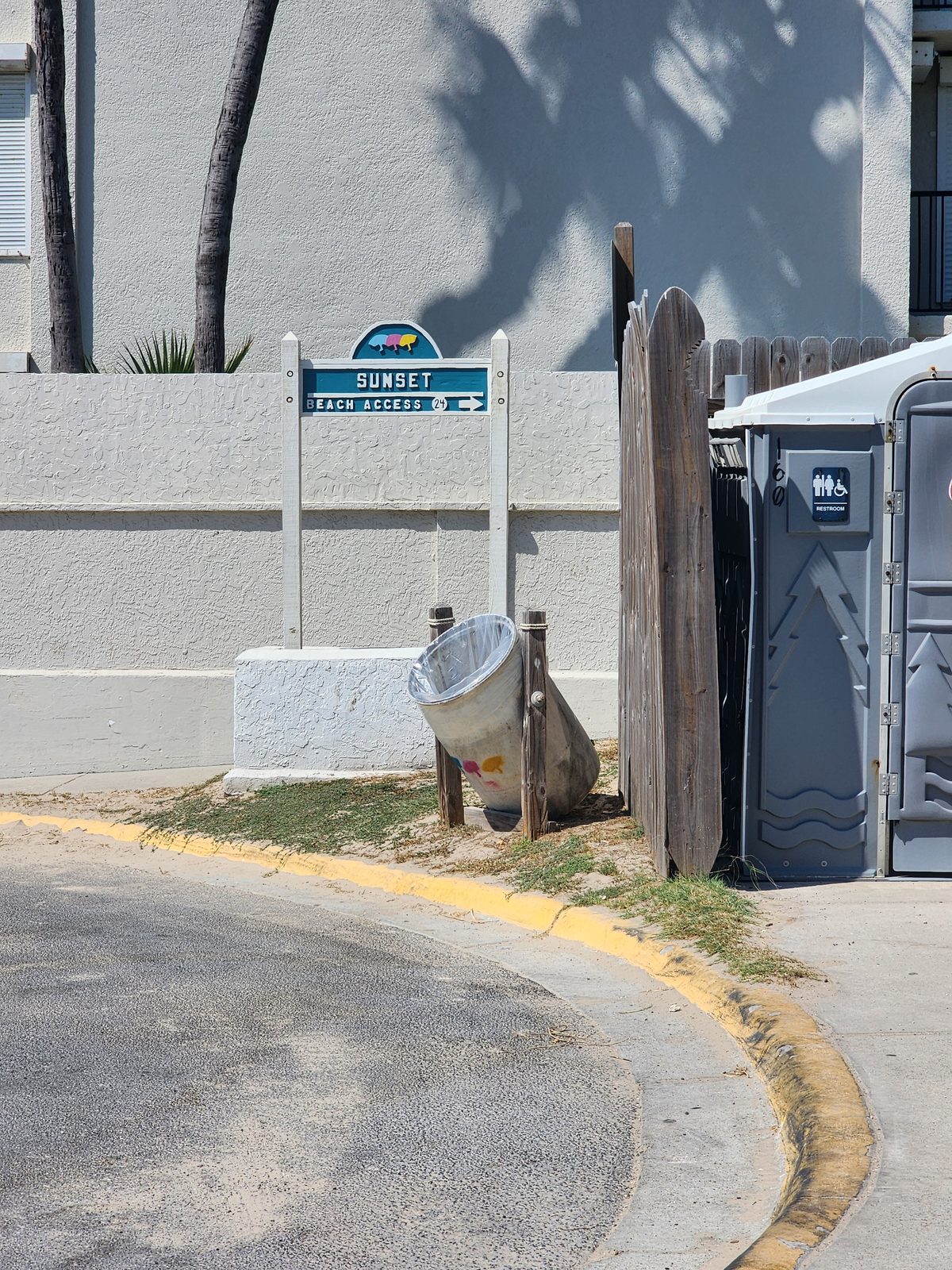 Teal Sunset Beach Access 24 sign with a right arrow on a stucco wall, beside a trash can, a wooden fence and a grey portable restroom.