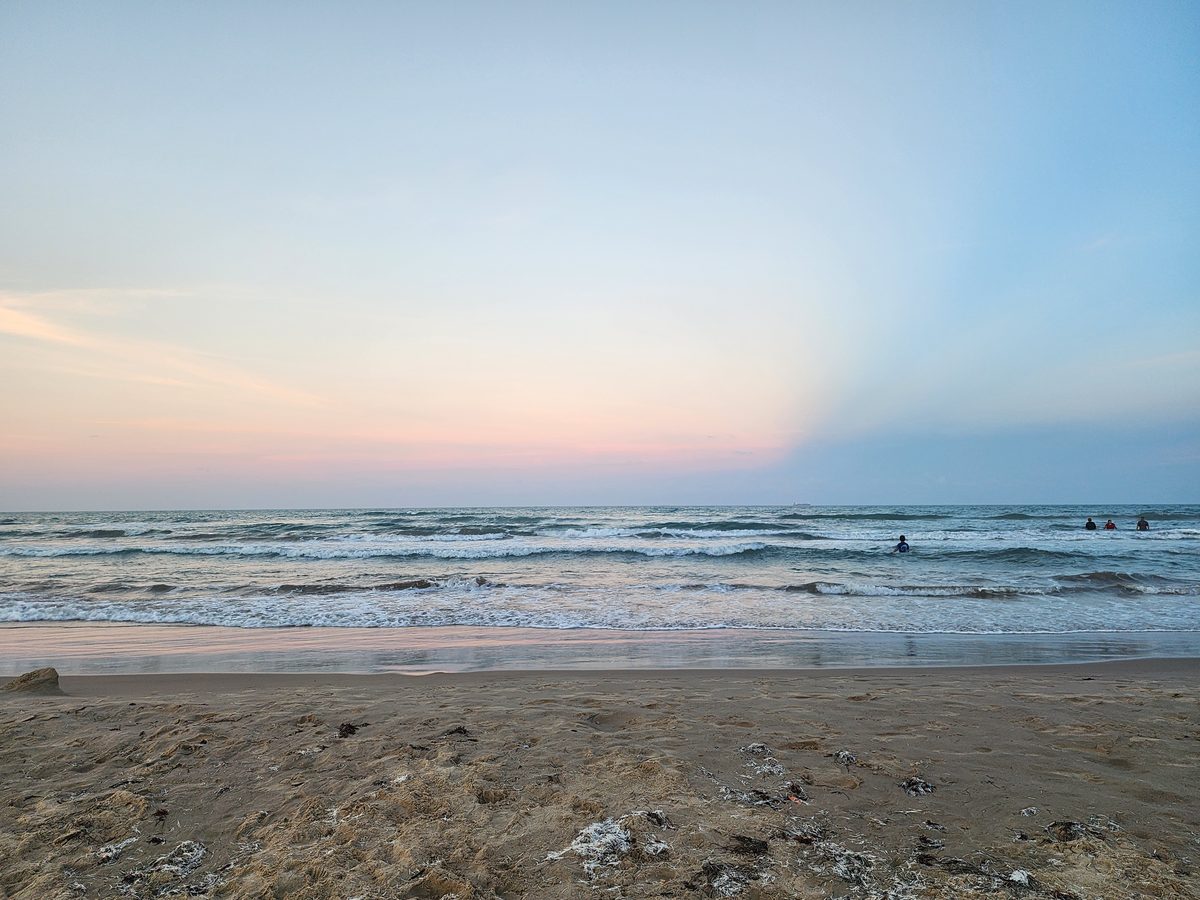 Pastel pink and blue sunset sky over Gulf waves, with a few distant swimmers in the surf and a sandy beach in the foreground.