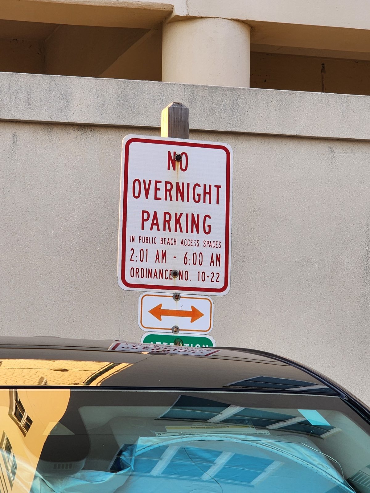 A red-and-white sign on a wooden post reads No Overnight Parking in Public Beach Access Spaces, 2:01 AM to 6:00 AM, above the roof of a parked car.