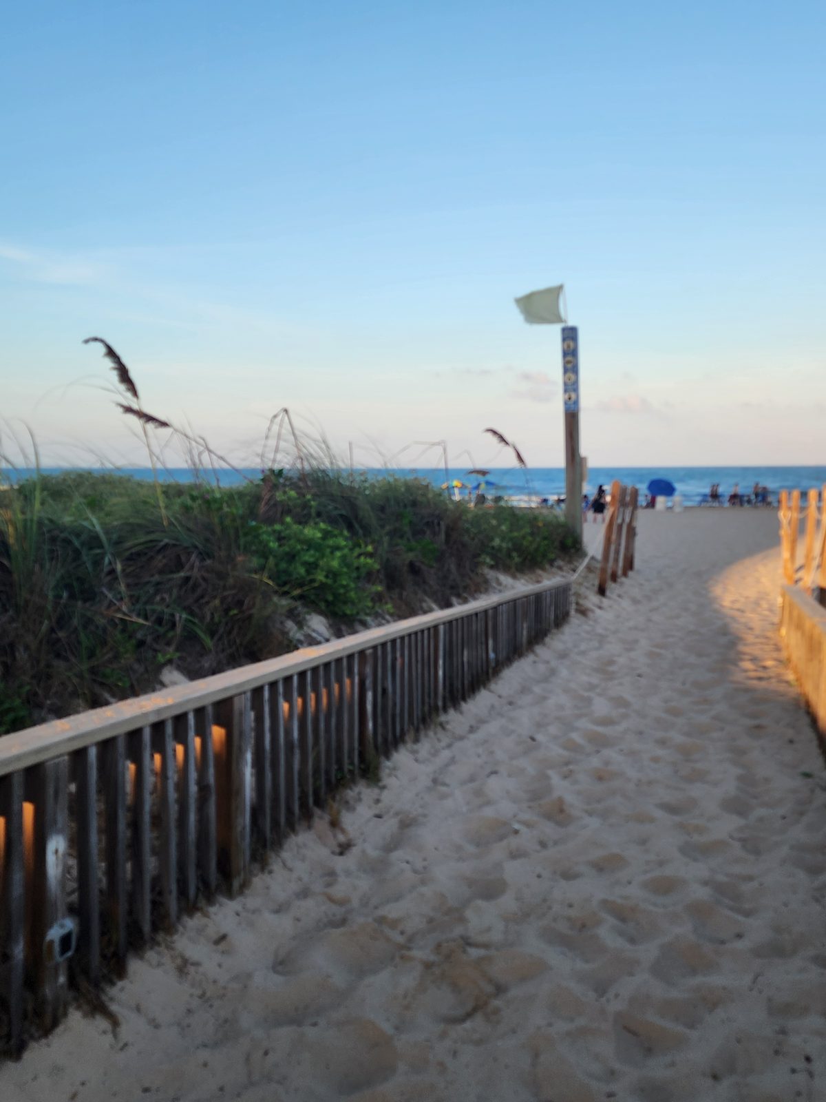 Sandy path between low wooden dune fences leading out to the Gulf beach, with a pale flag on a signpost and distant beachgoers near the water.