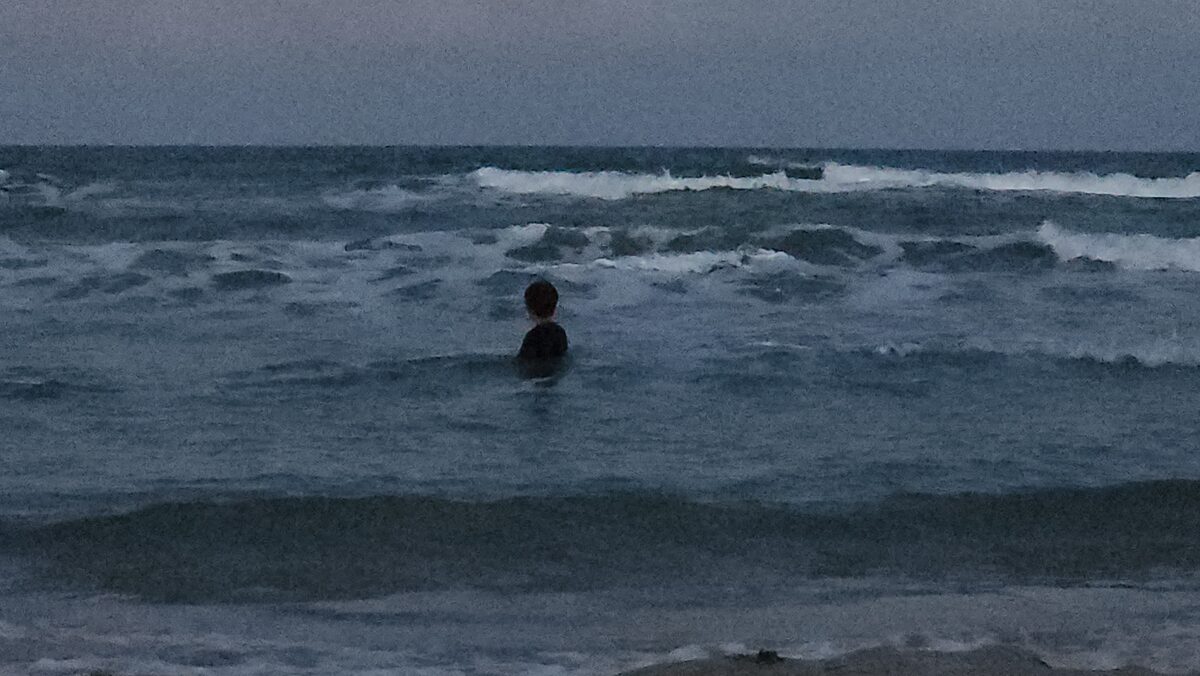 A child seen from behind, chest-deep in choppy grey Gulf surf at dusk, facing rows of white breakers under a flat grey sky.