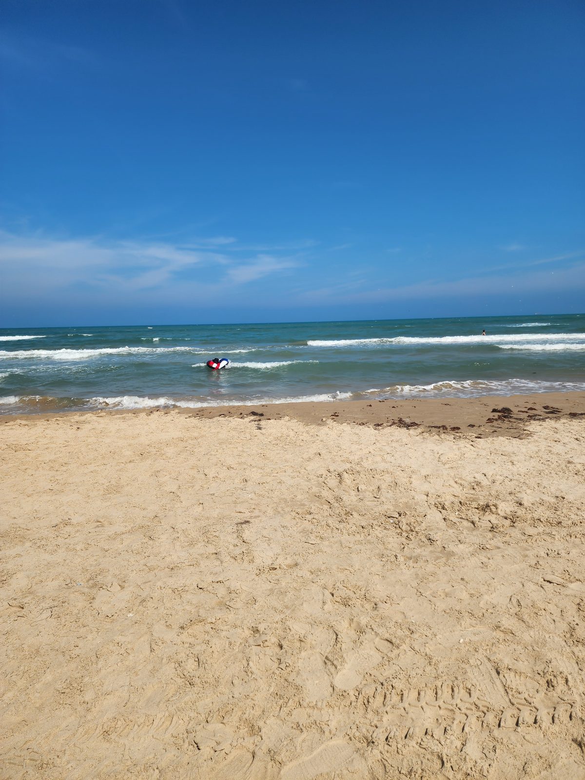 Across wide open sand, a person on a red, white and blue inflatable in the Gulf surf, with a gull at the waterline and another swimmer farther out.