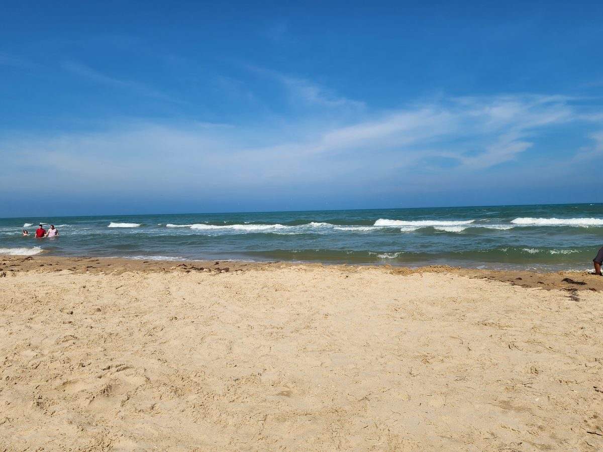 Distant visitors wade in the Gulf beside a sandy beach under a blue sky.
