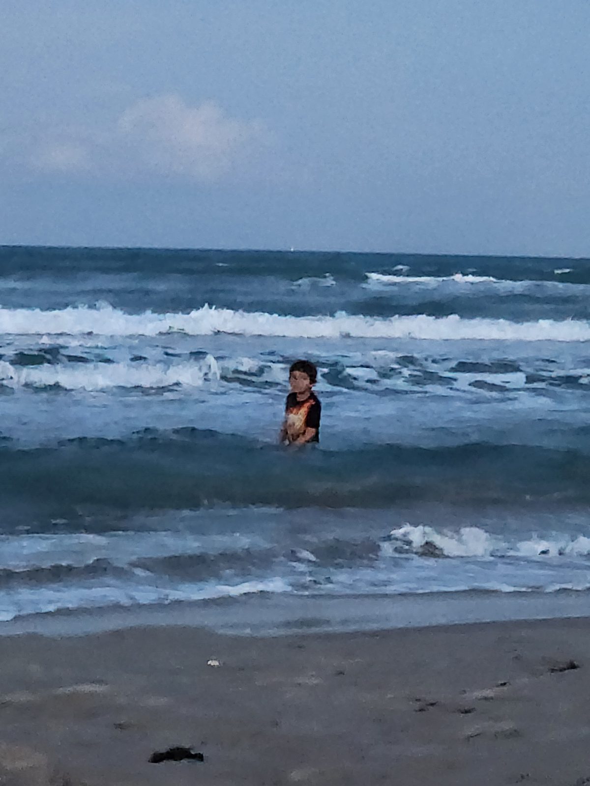 A child stands in the breaking Gulf surf at dusk, with lines of white breakers behind and wet sand in the foreground.