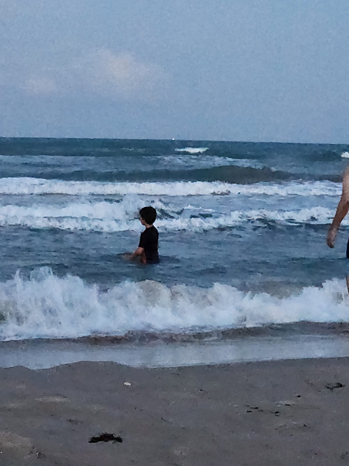 A child chest-deep in choppy Gulf surf at dusk, rows of breaking waves behind under a plain twilight sky.