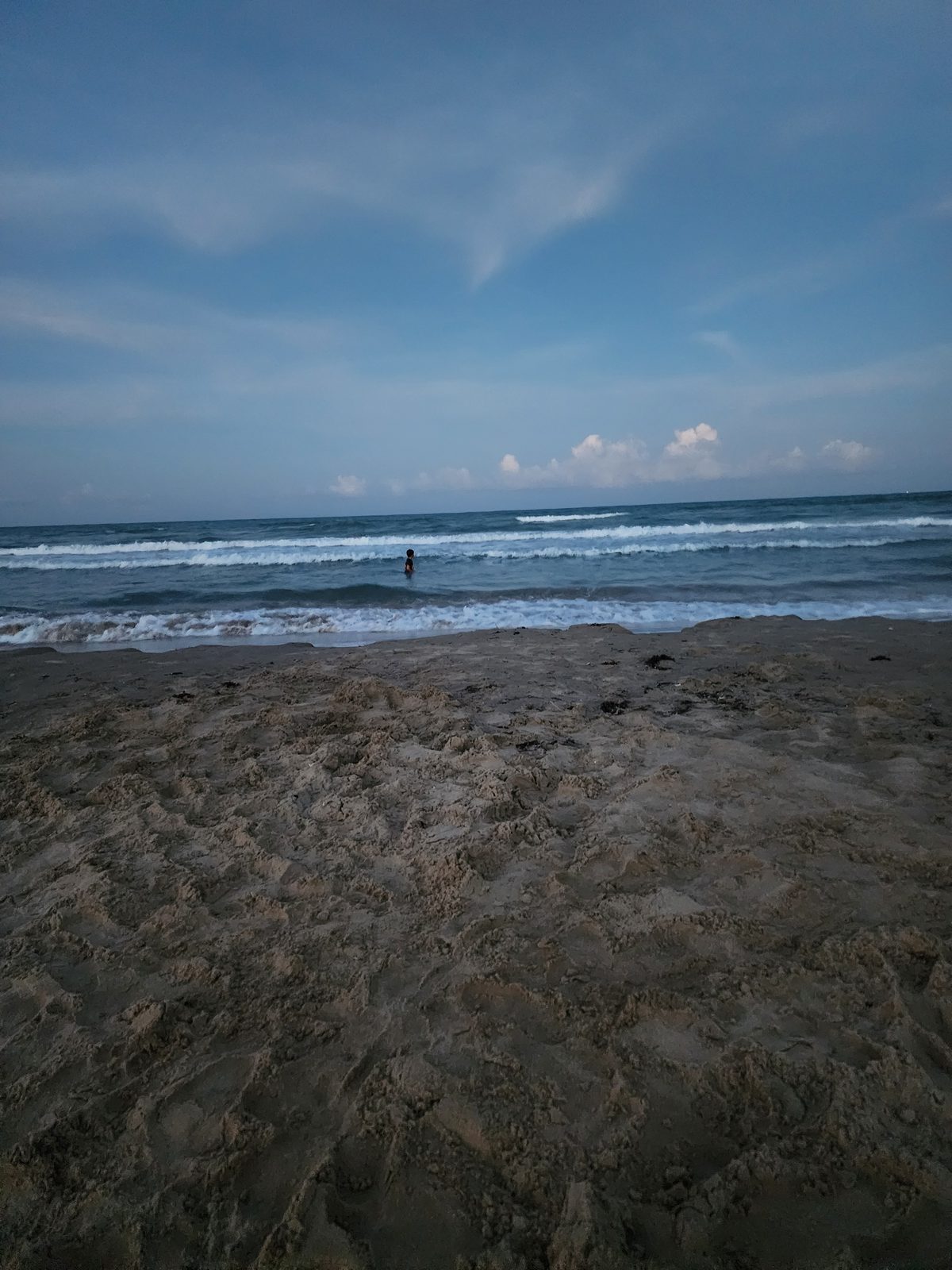 A lone person wades in the Gulf surf beyond a wide, footprint-covered sandy beach under a dusky blue evening sky.