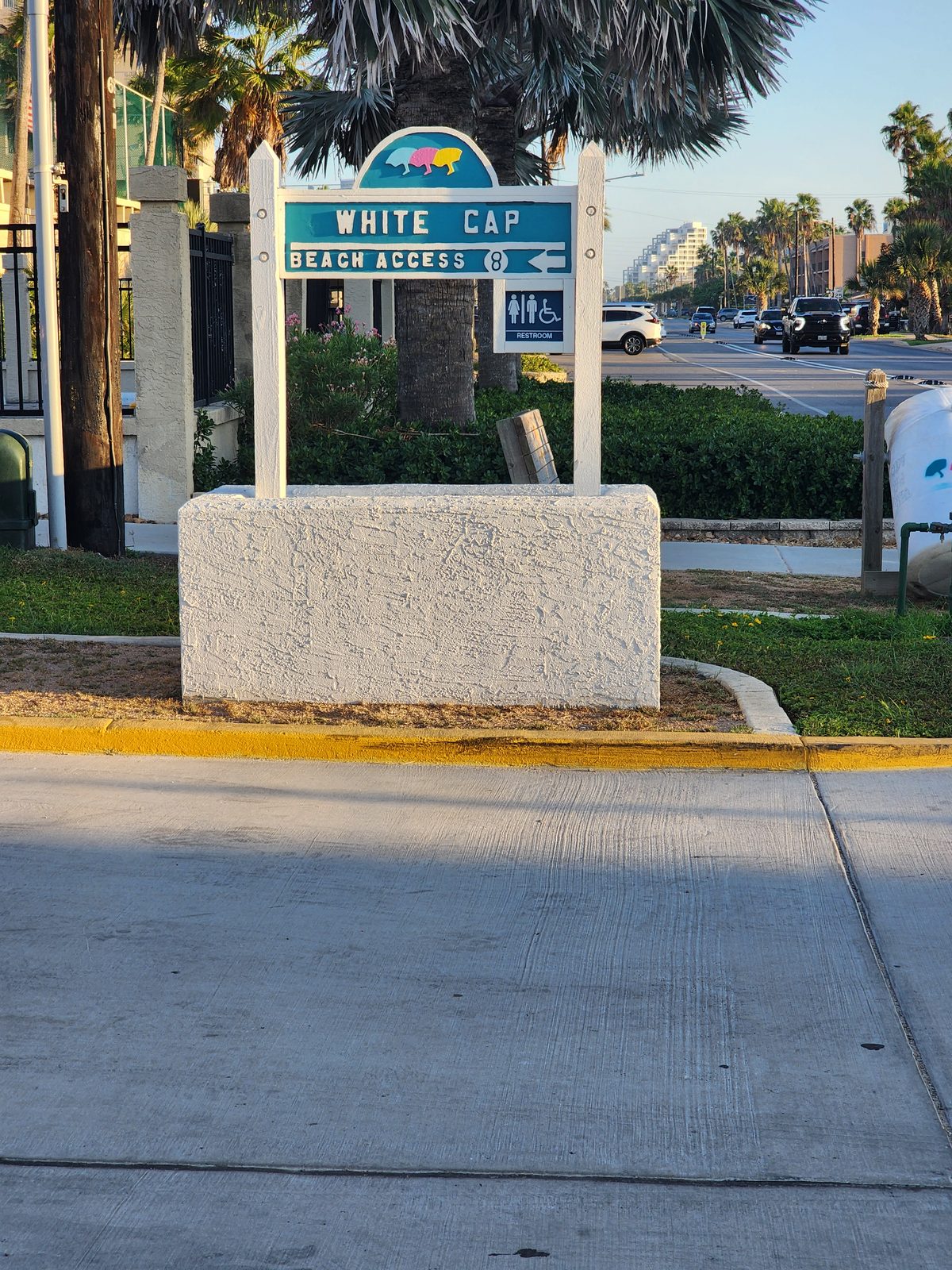A teal White Cap Beach Access 8 sign with a left arrow and restroom symbol sits on a white stucco base near palms and a road.