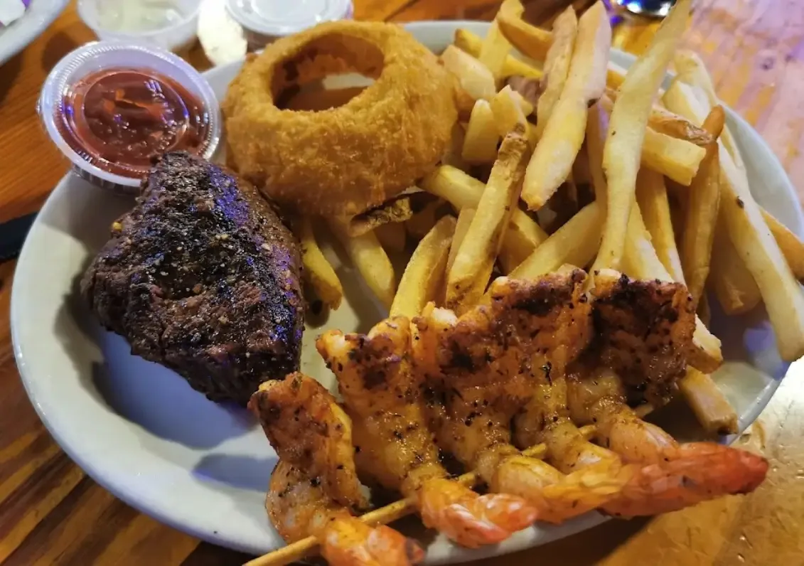 A steak with a skewer of grilled shrimp, an onion ring and fries