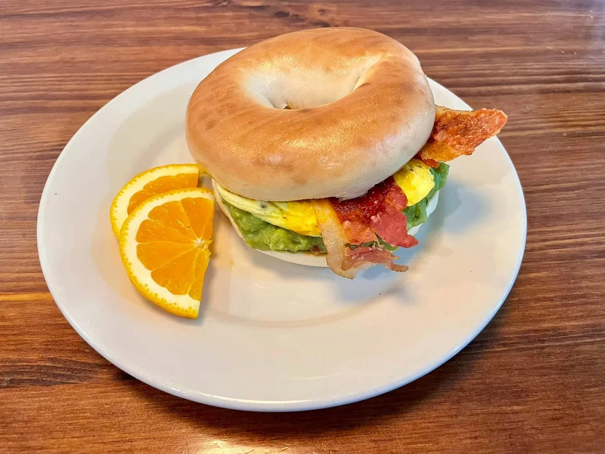 A bagel sandwich with egg, bacon and avocado, with orange slices