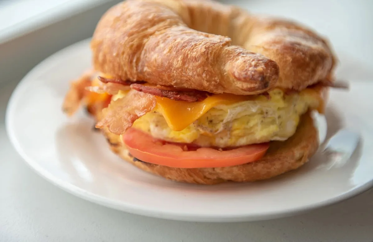 A croissant breakfast sandwich with egg, bacon, cheddar and tomato on a white plate
