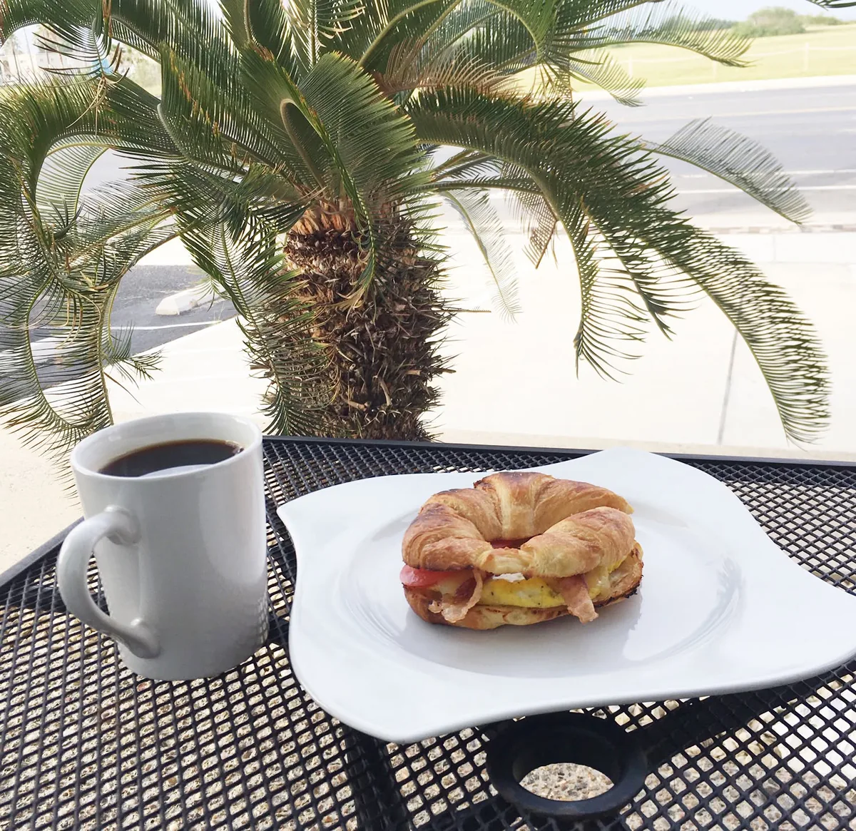 A mug of coffee and a croissant sandwich on a mesh patio table under a palm