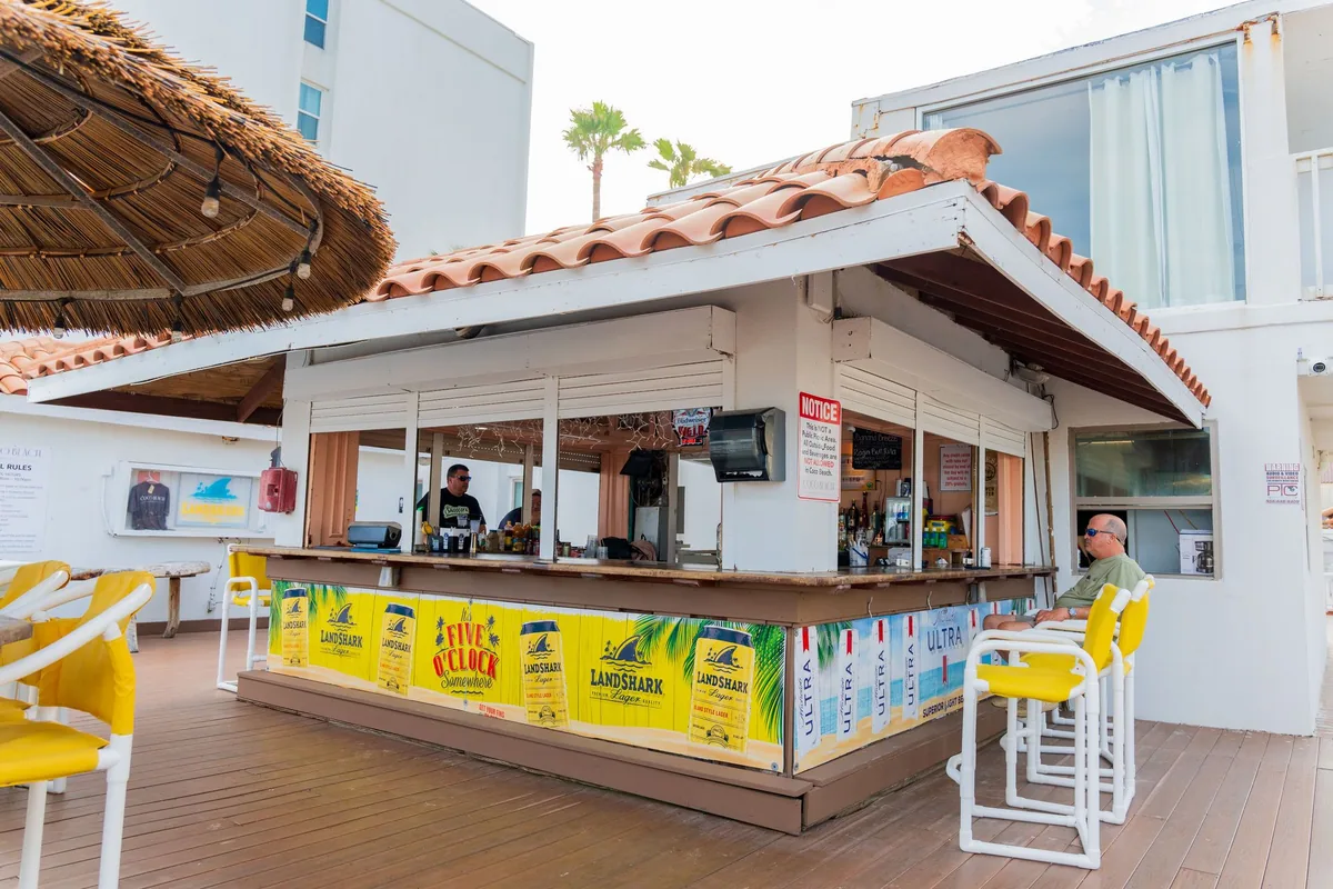 The Coco Beach Bar, a white bar hut with a tile roof and yellow bar stools, next to a thatched umbrella